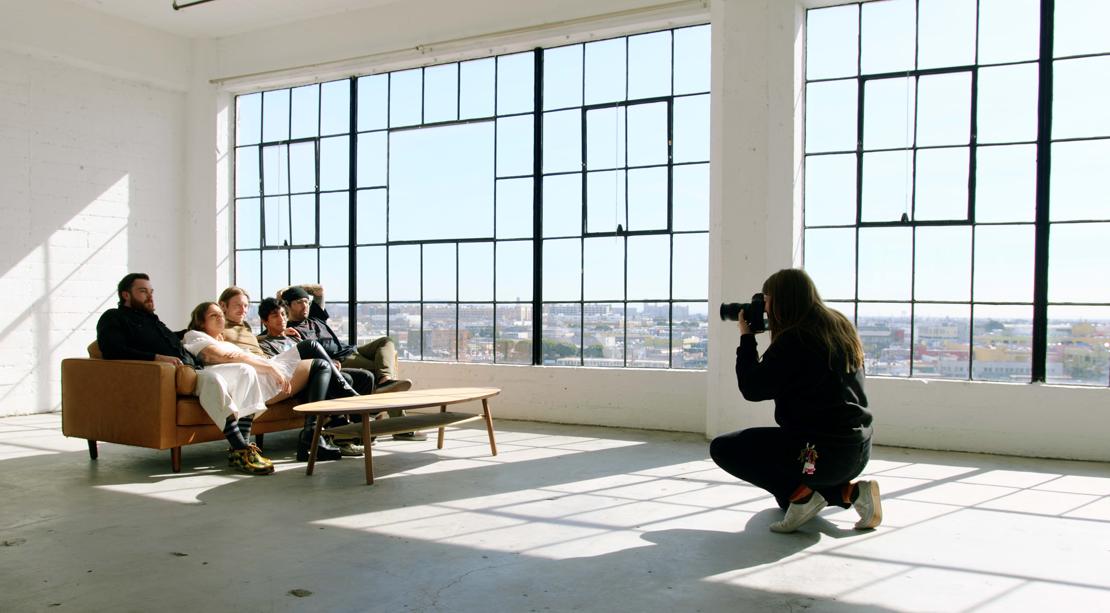 A photographer captures a group of four people sitting on a couch in a bright, spacious room with large windows.
