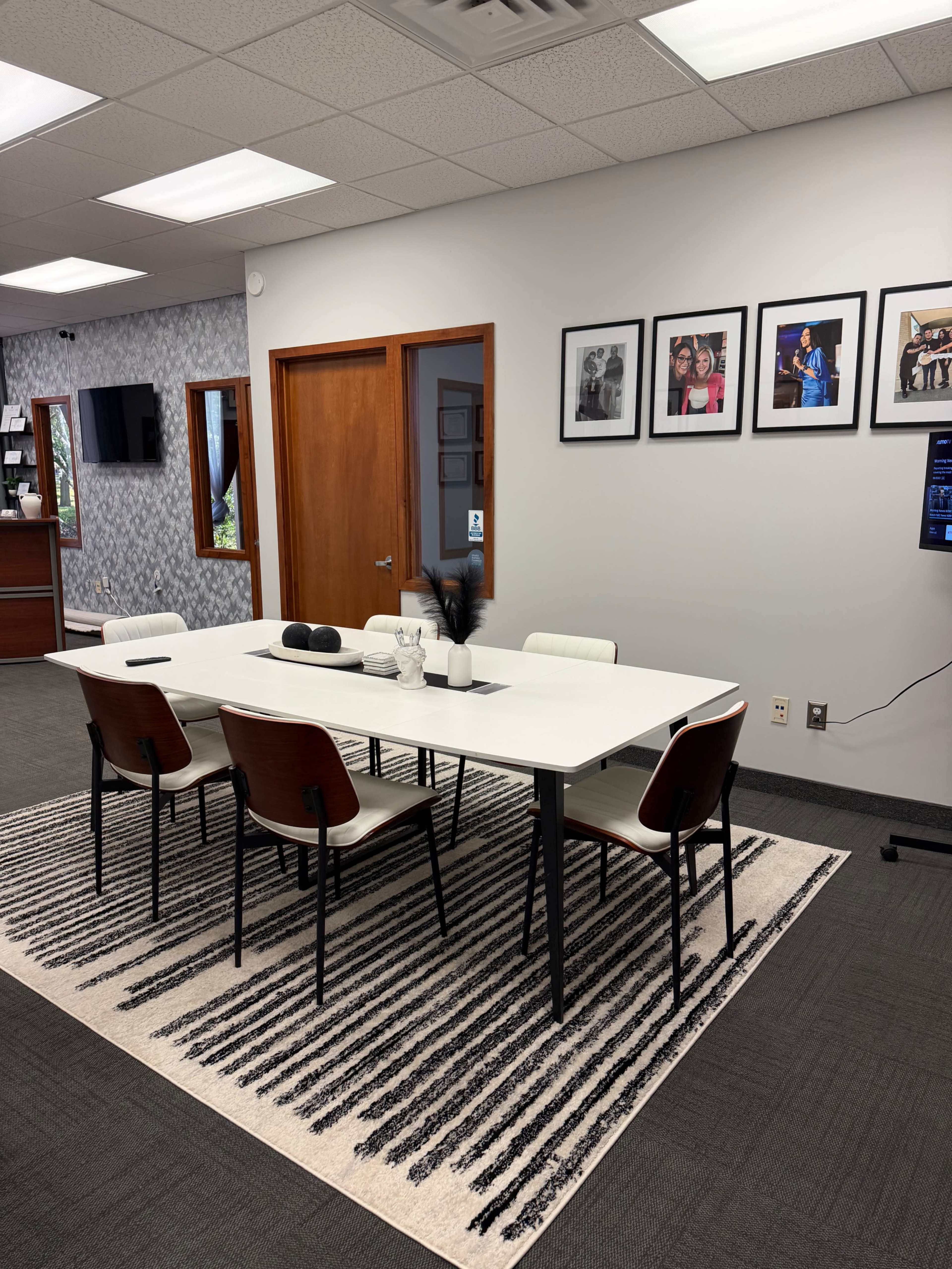 The image shows a modern conference room featuring a long white table surrounded by six chairs, a patterned rug on the floor, and framed photographs on the wall.