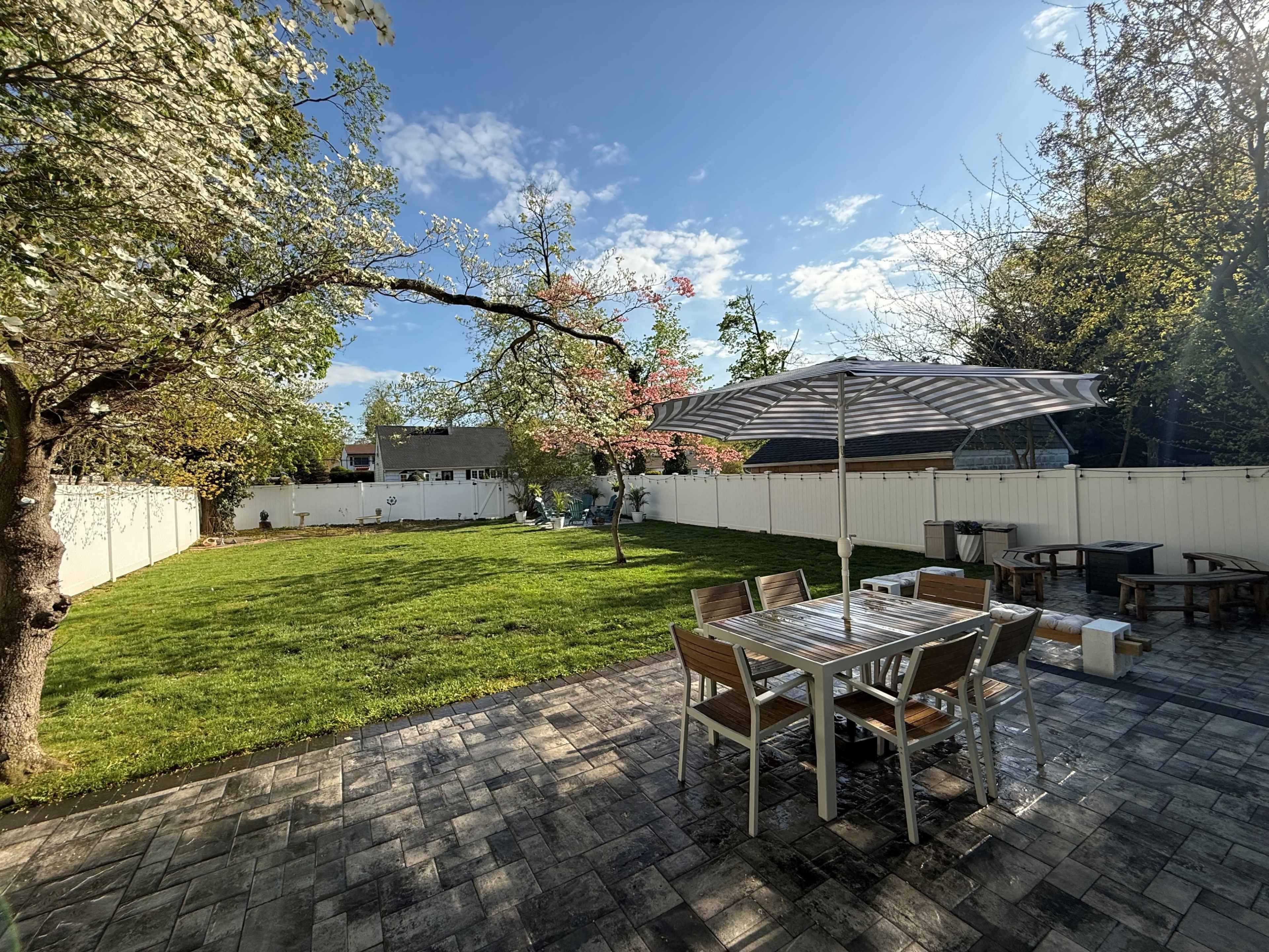 The image shows a spacious backyard with a grassy area, a wooden dining table with chairs, an umbrella, and a seating arrangement surrounded by trees and a white fence.