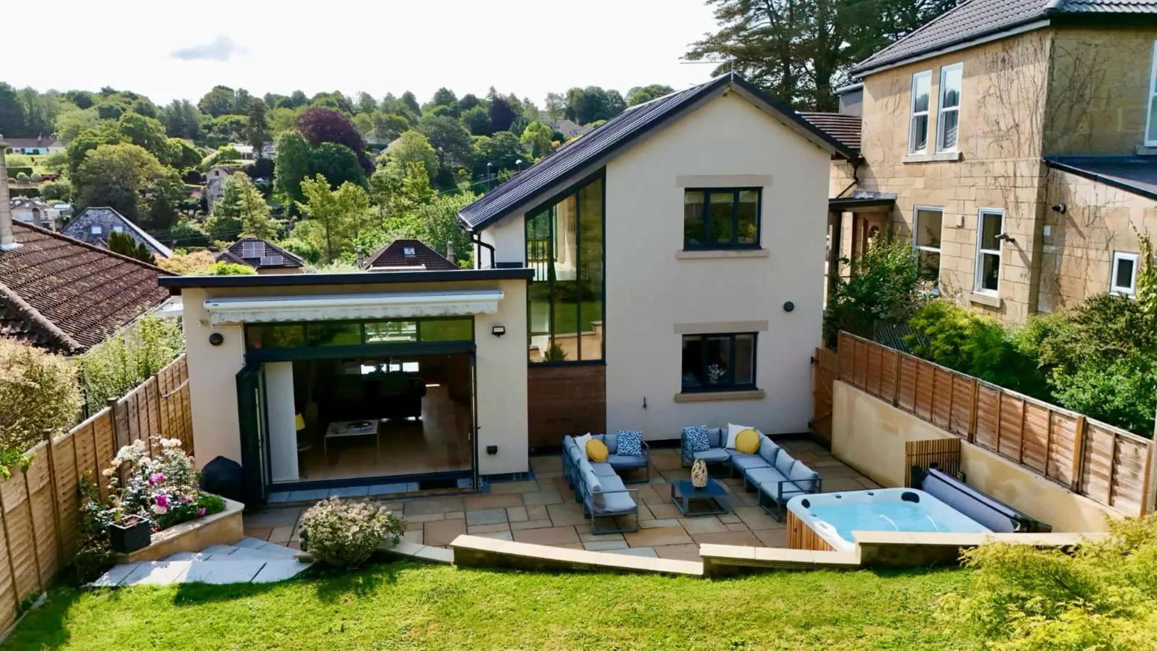 The image shows a modern house with large windows, a patio area featuring outdoor seating and a hot tub, surrounded by greenery in a residential neighborhood.
