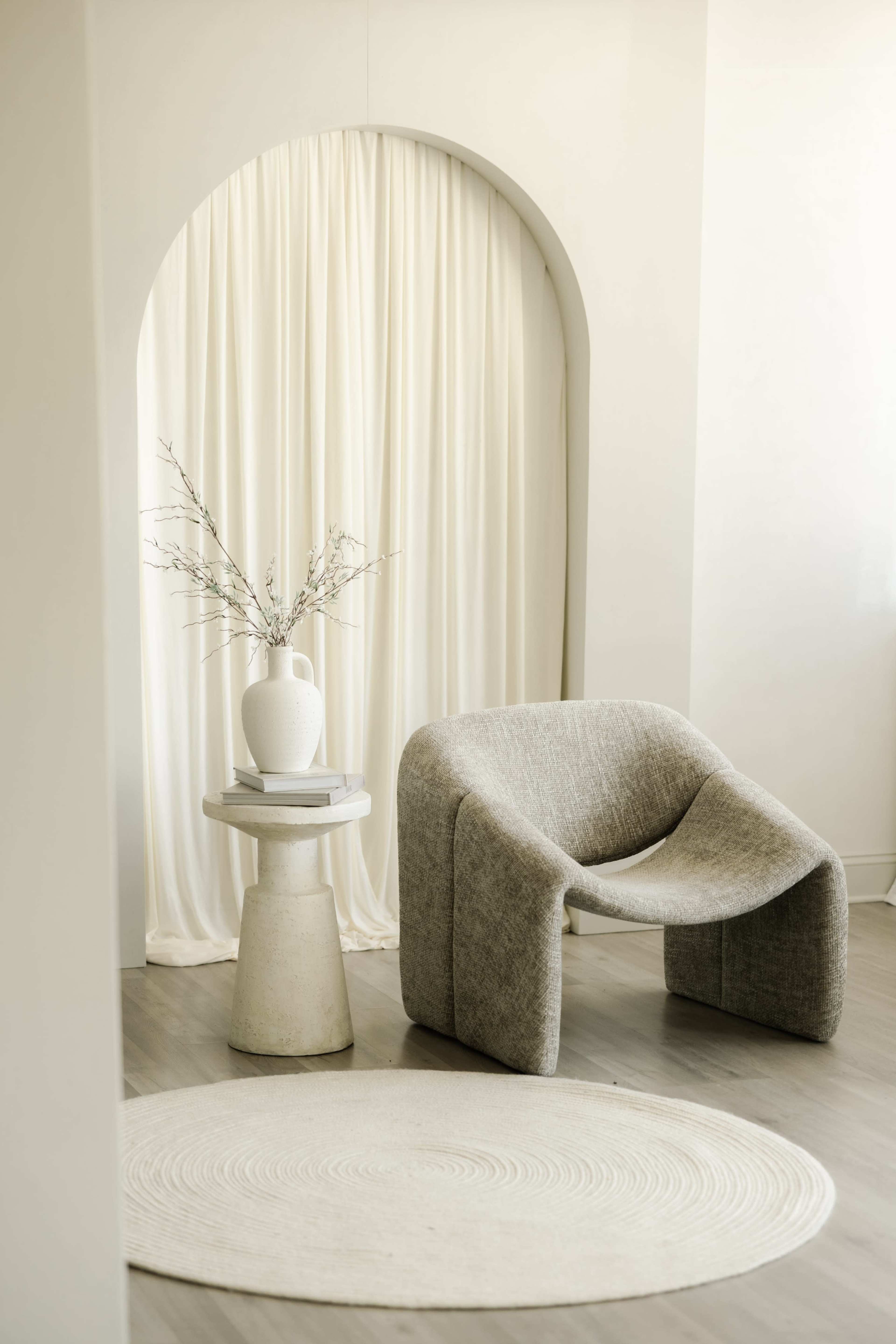 The scene features a minimalist interior with a gray upholstered chair, a round rug on the floor, a cylindrical table, and a vase with branches beside a sheer curtain.