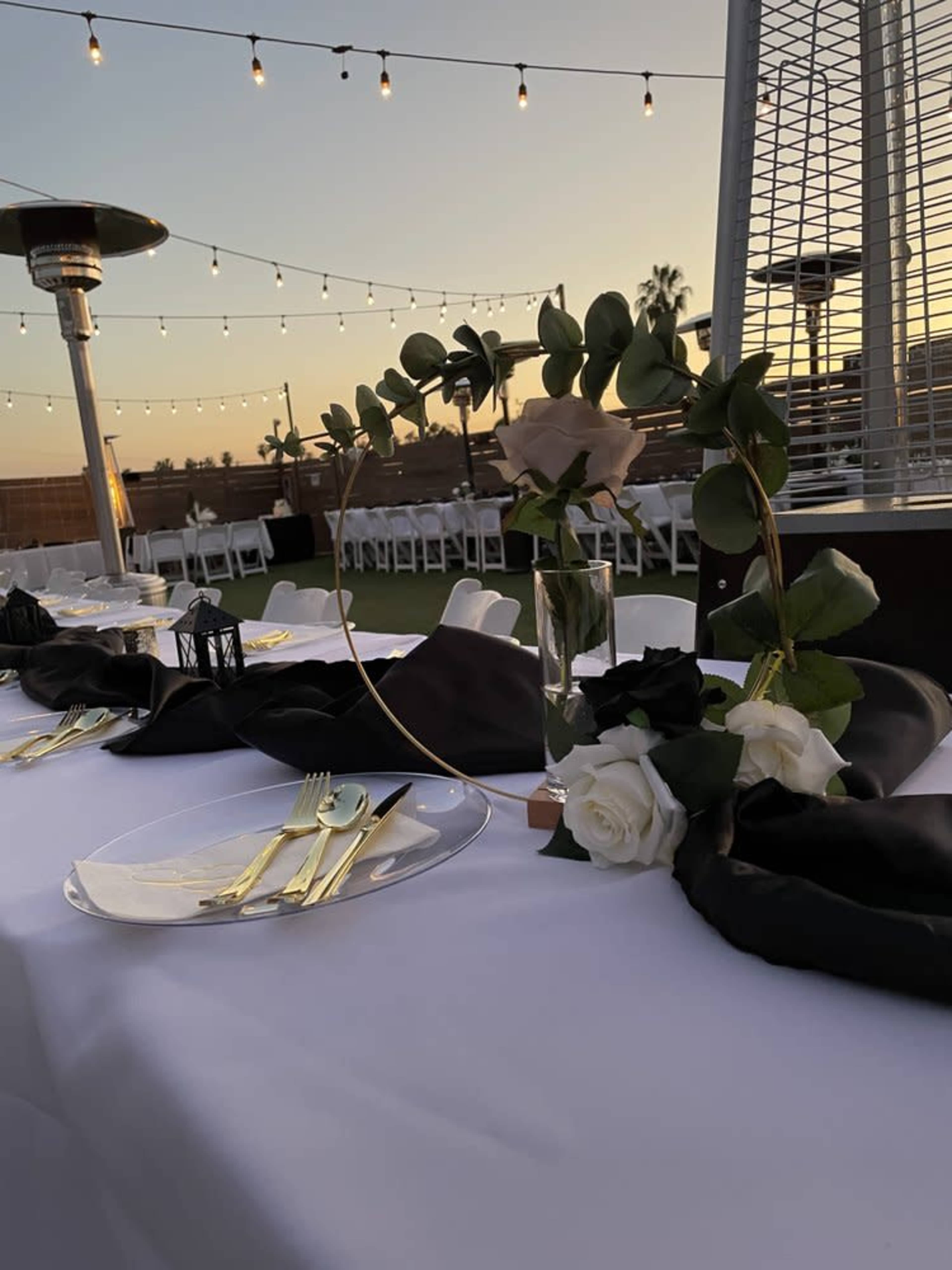 A elegantly set table for an outdoor event features gold flatware and floral centerpieces, illuminated by string lights against a sunset backdrop.