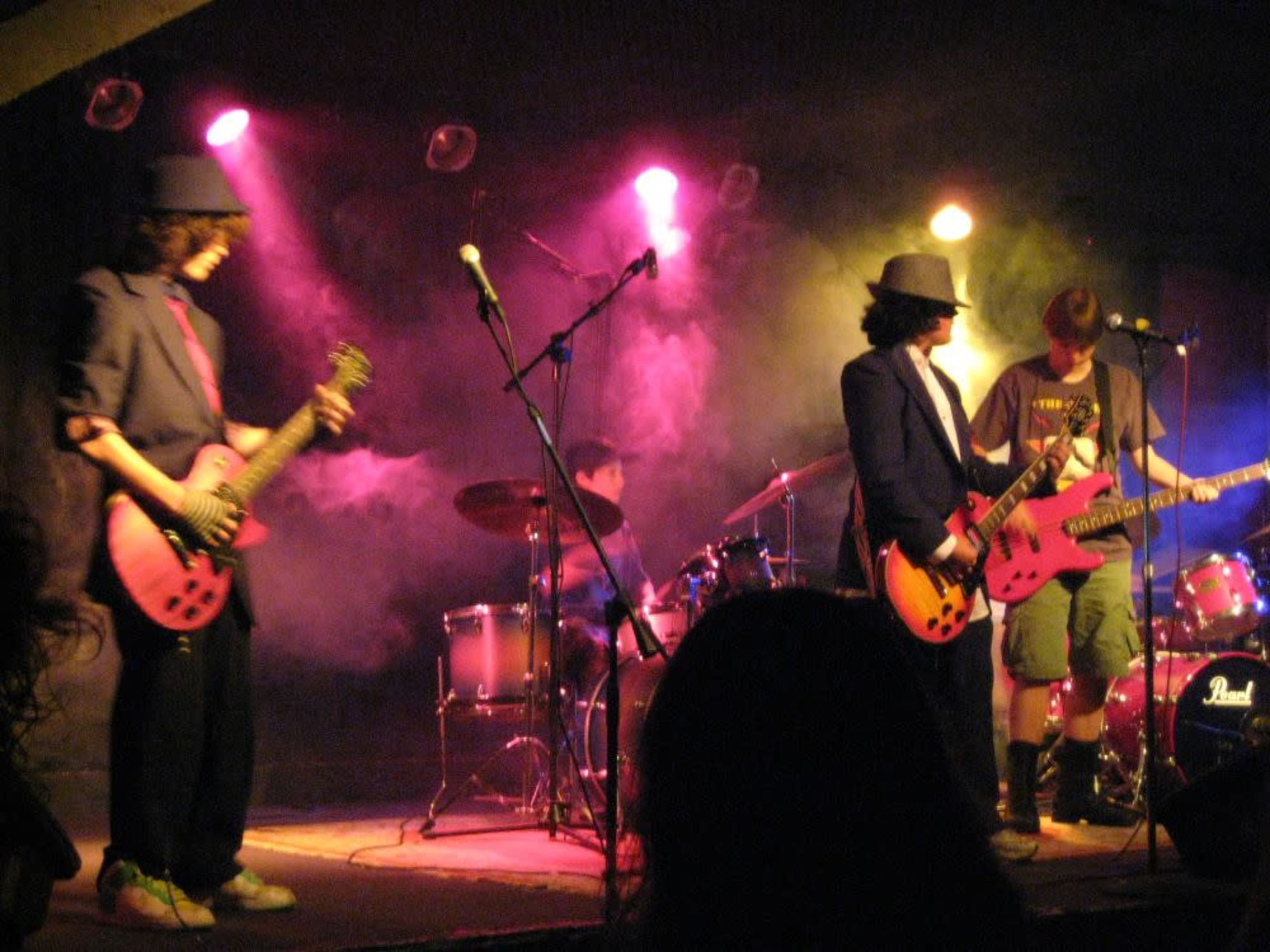 A band performs on stage with electric guitars and a drum set, surrounded by colorful stage lights and fog.