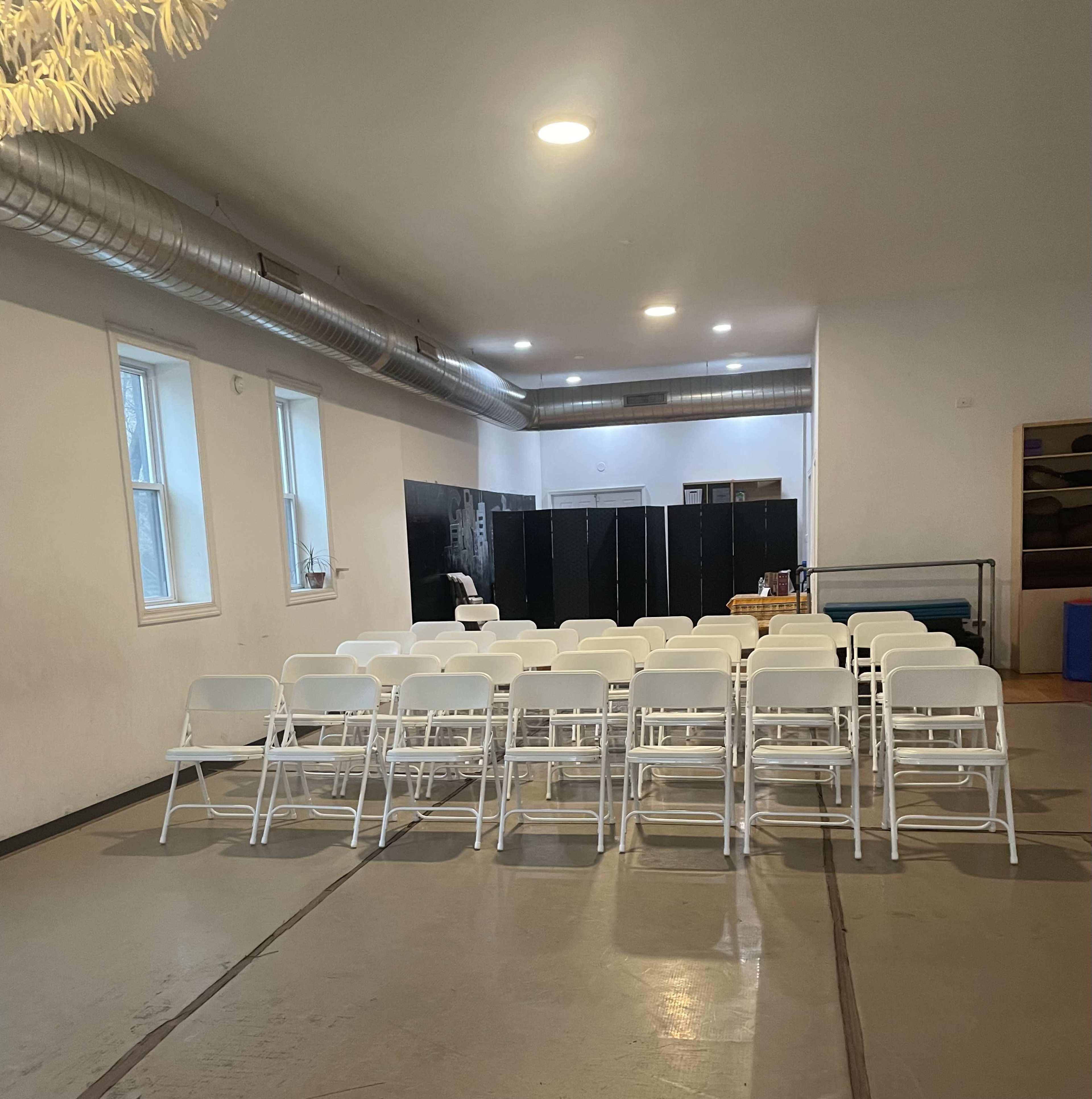 The image shows a spacious room with rows of white folding chairs facing a black partition and various items in the background.