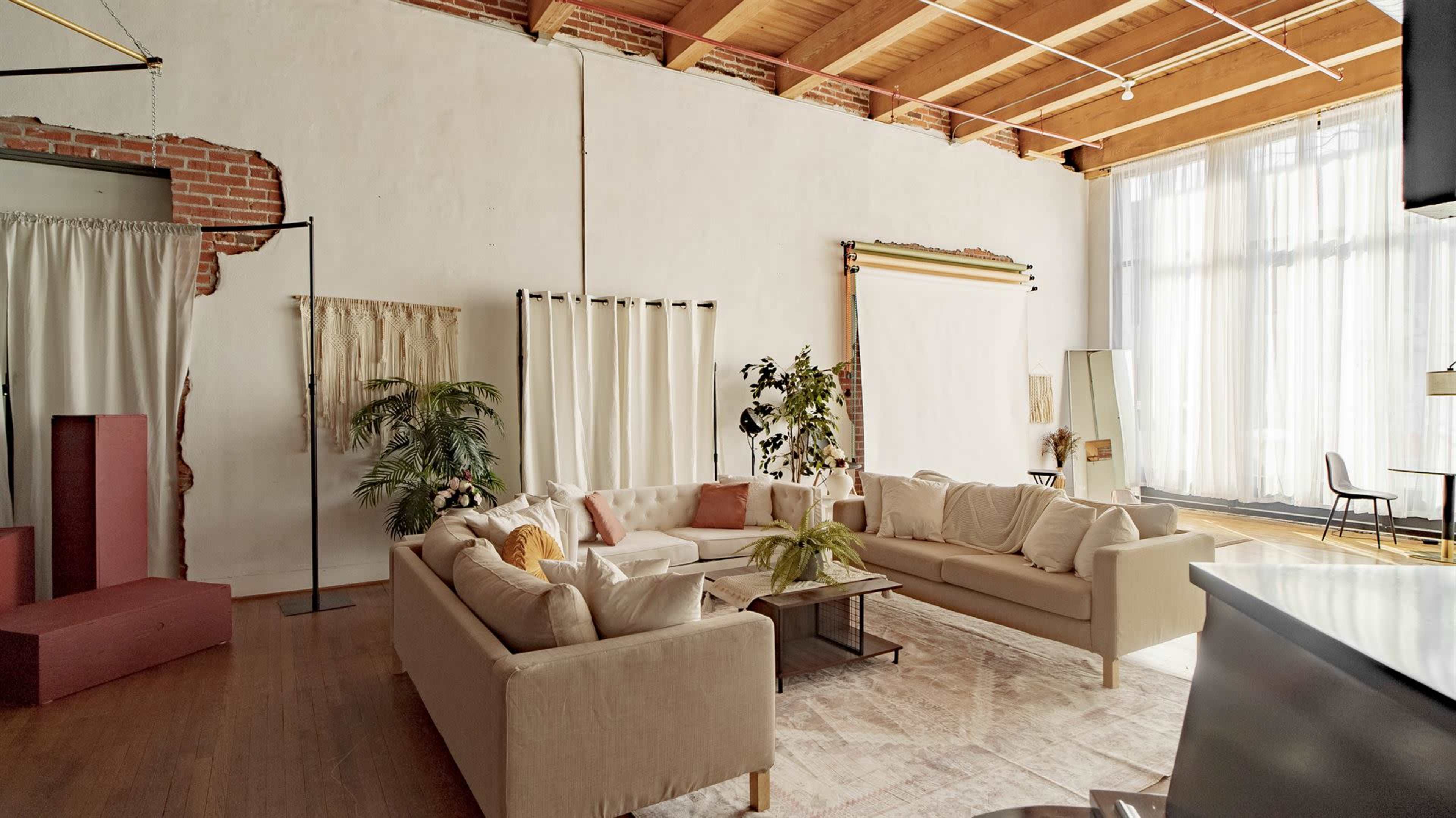The image shows a spacious, well-lit living area featuring a large beige sectional sofa, a coffee table, and decorative plants against a backdrop of exposed brick and wooden beams.