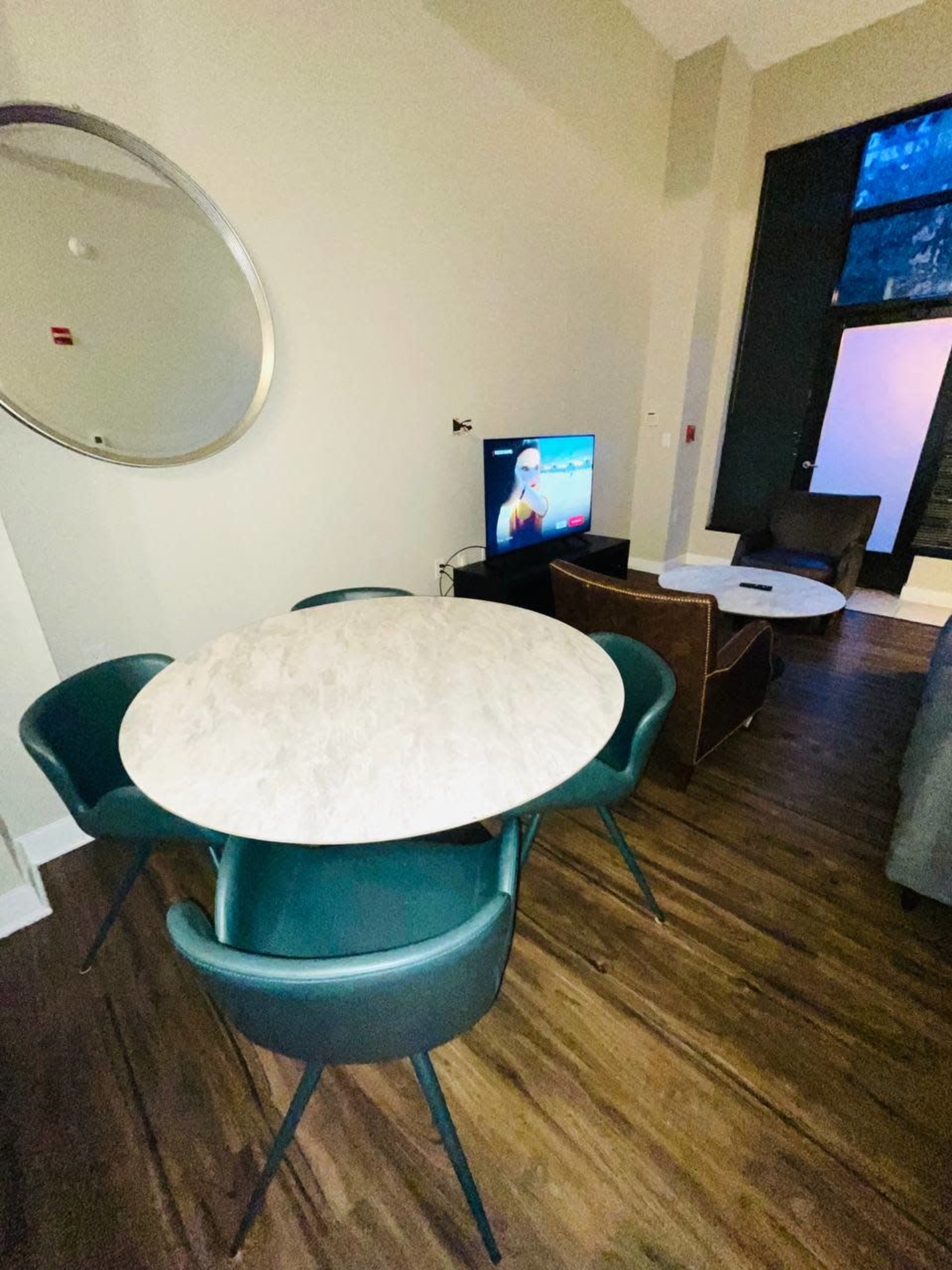 A modern living space featuring a round marble table with four green chairs, a brown armchair, and a television displaying a video in a well-lit room.