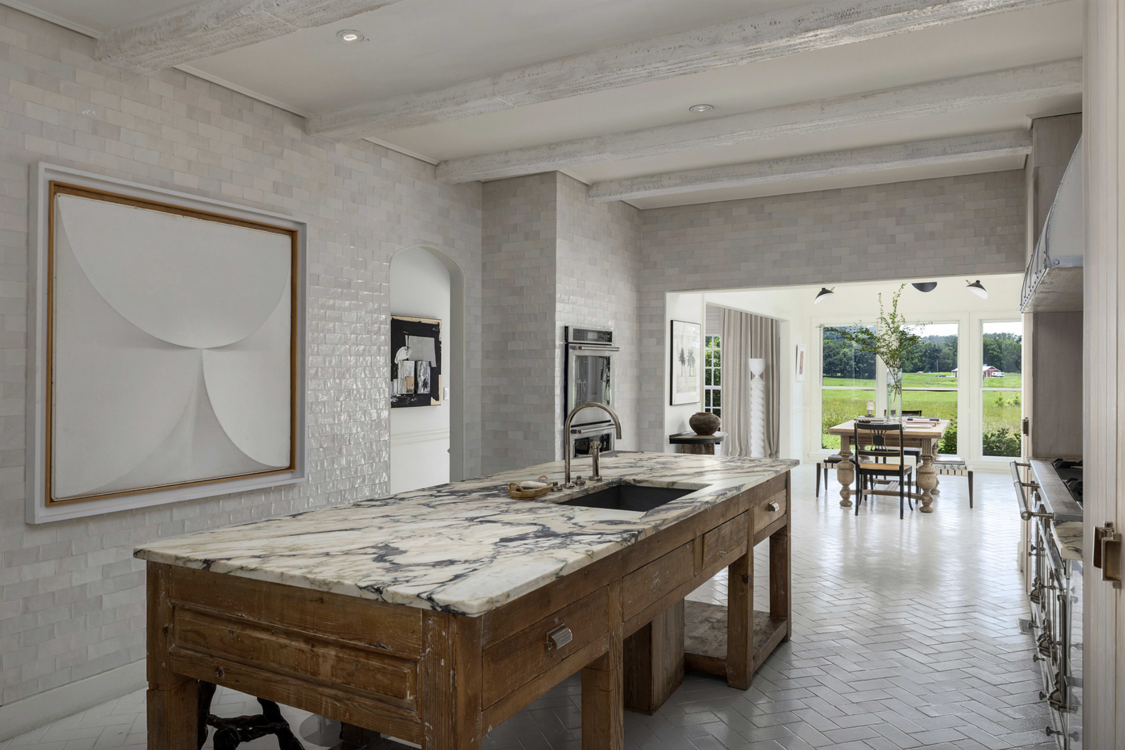 The image shows a modern kitchen featuring a large marble-topped island with wooden cabinetry, light-colored tiled walls, and a view of an outdoor dining area through large windows.