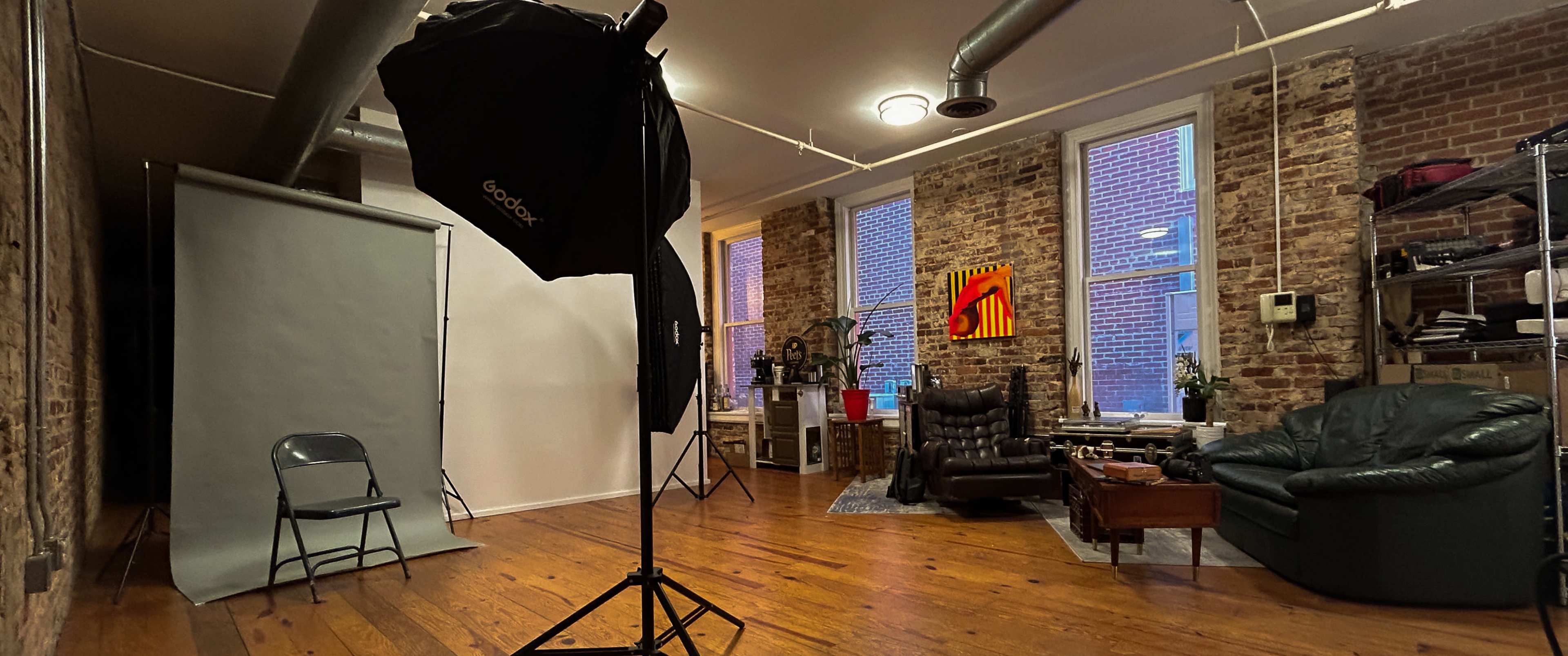 A photography studio features a gray backdrop, a softbox light, a single chair, and a cozy seating area with a green couch and wooden furniture.