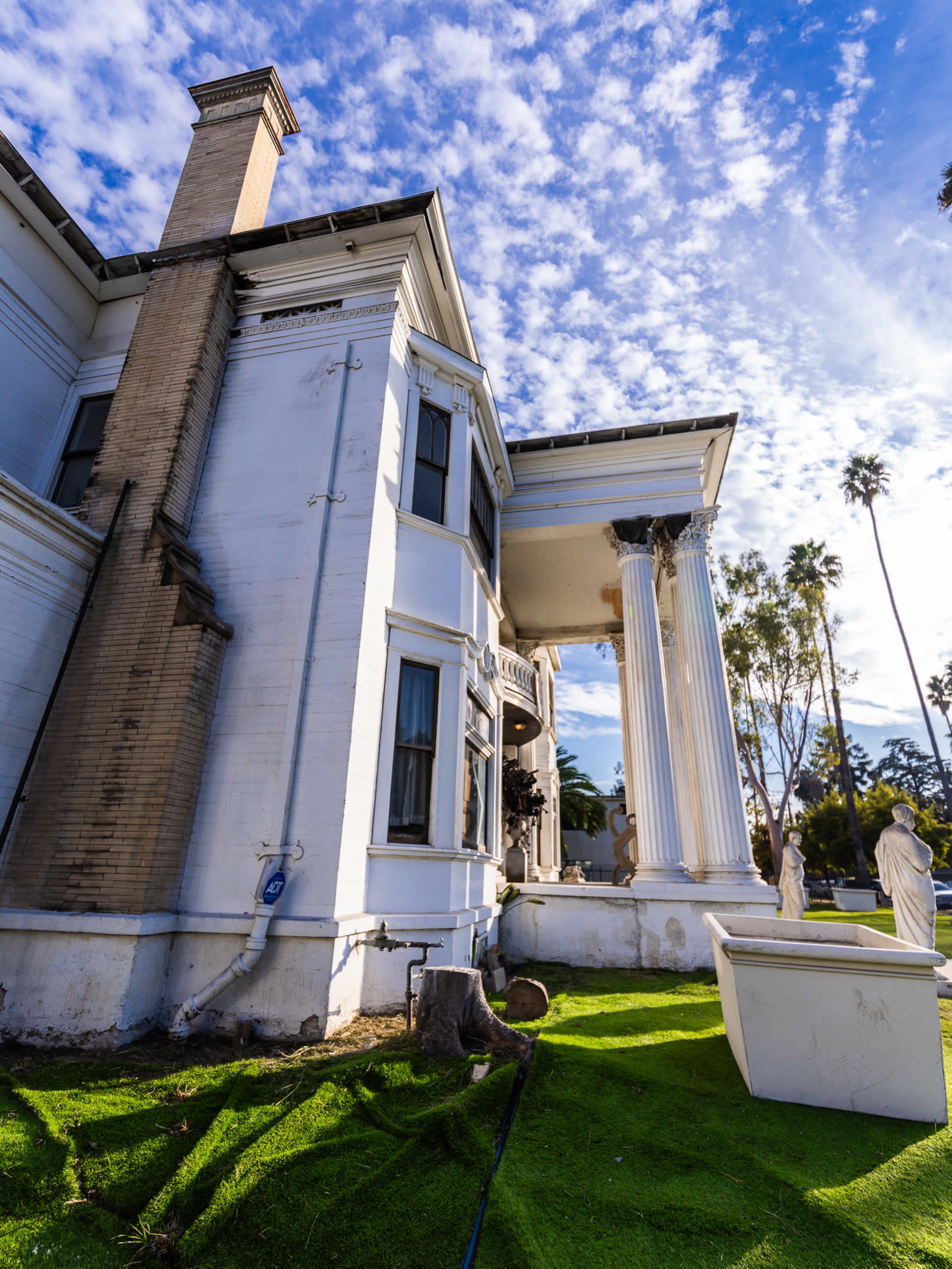 A large white mansion with decorative columns and statues in the garden, under a blue sky with scattered clouds.