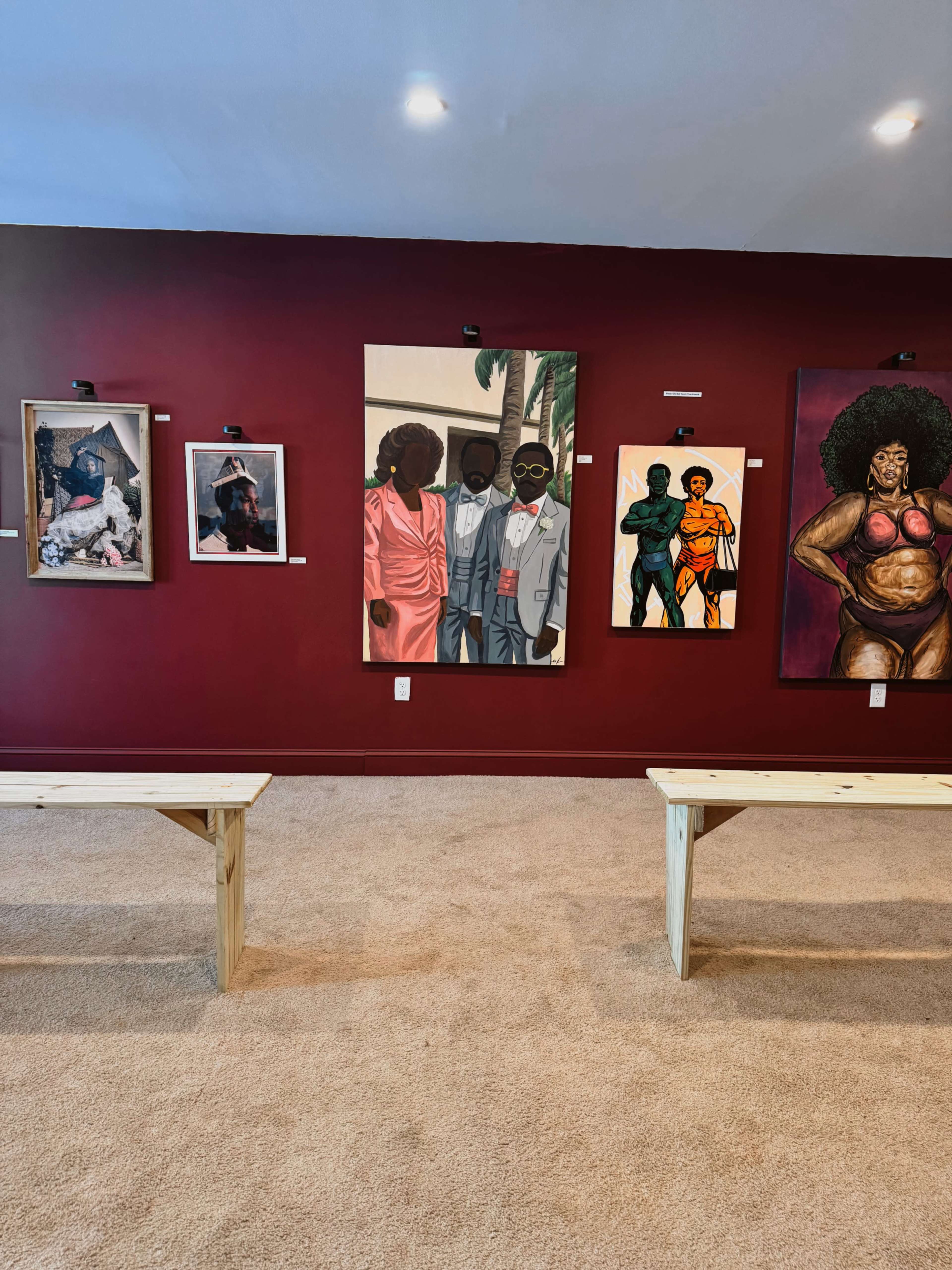 The image shows an art gallery with a red wall displaying various framed paintings and photographs, along with two wooden benches in the foreground.