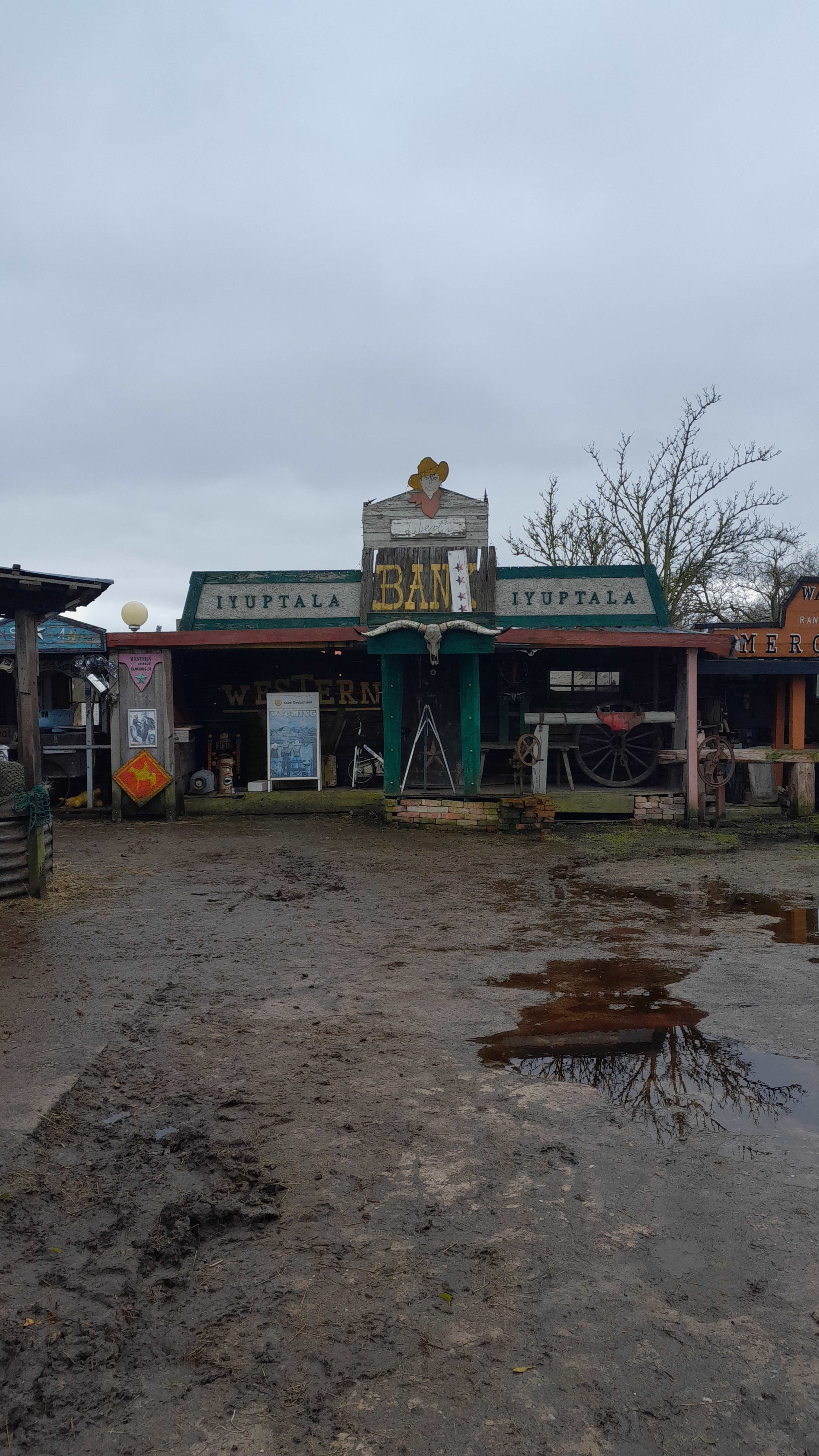 The image shows a rustic storefront labeled "BAN" surrounded by muddy ground, with old western-themed decorations and structures in the background.