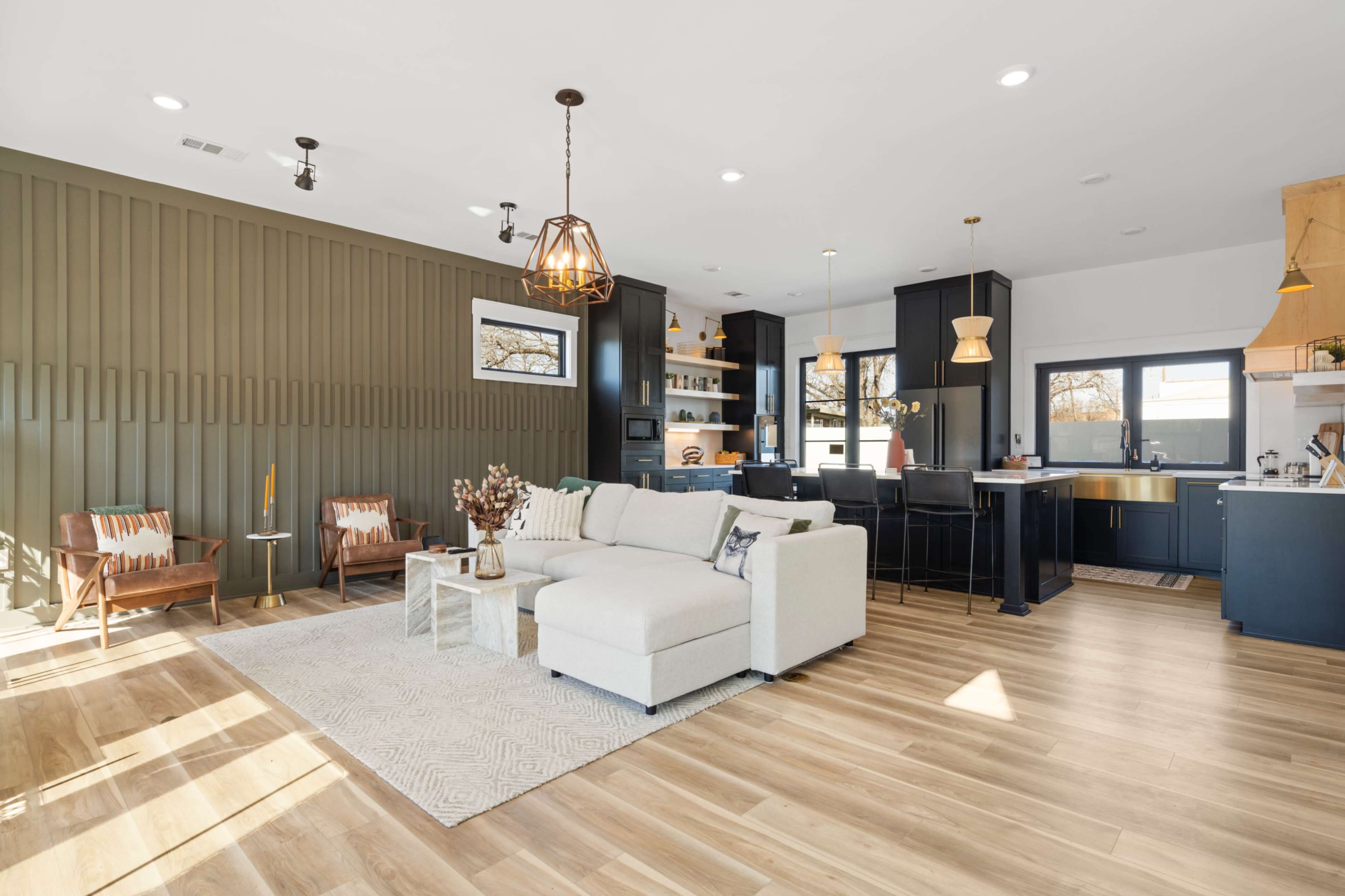 A modern living room features a light sectional sofa, wooden accents, and a dark kitchen area with stylish pendant lighting.