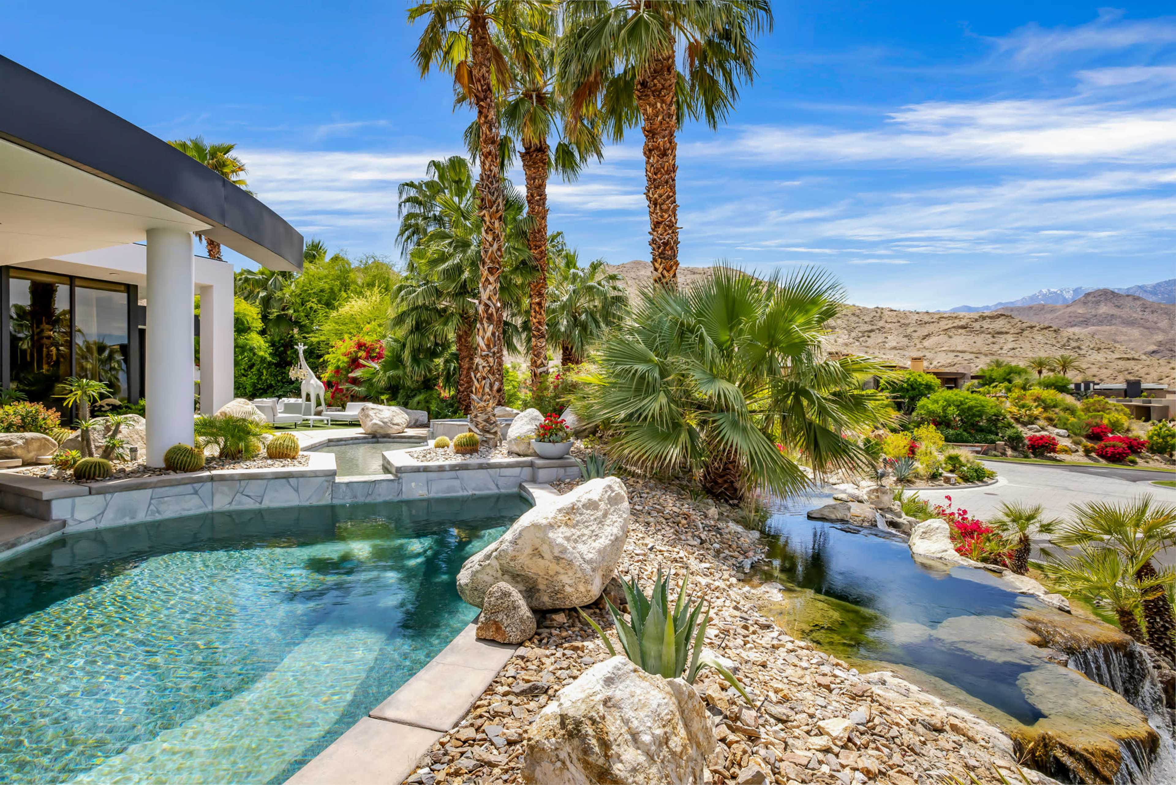The image shows a modern villa beside a pool, surrounded by palm trees, boulders, and lush greenery, with mountains in the background.