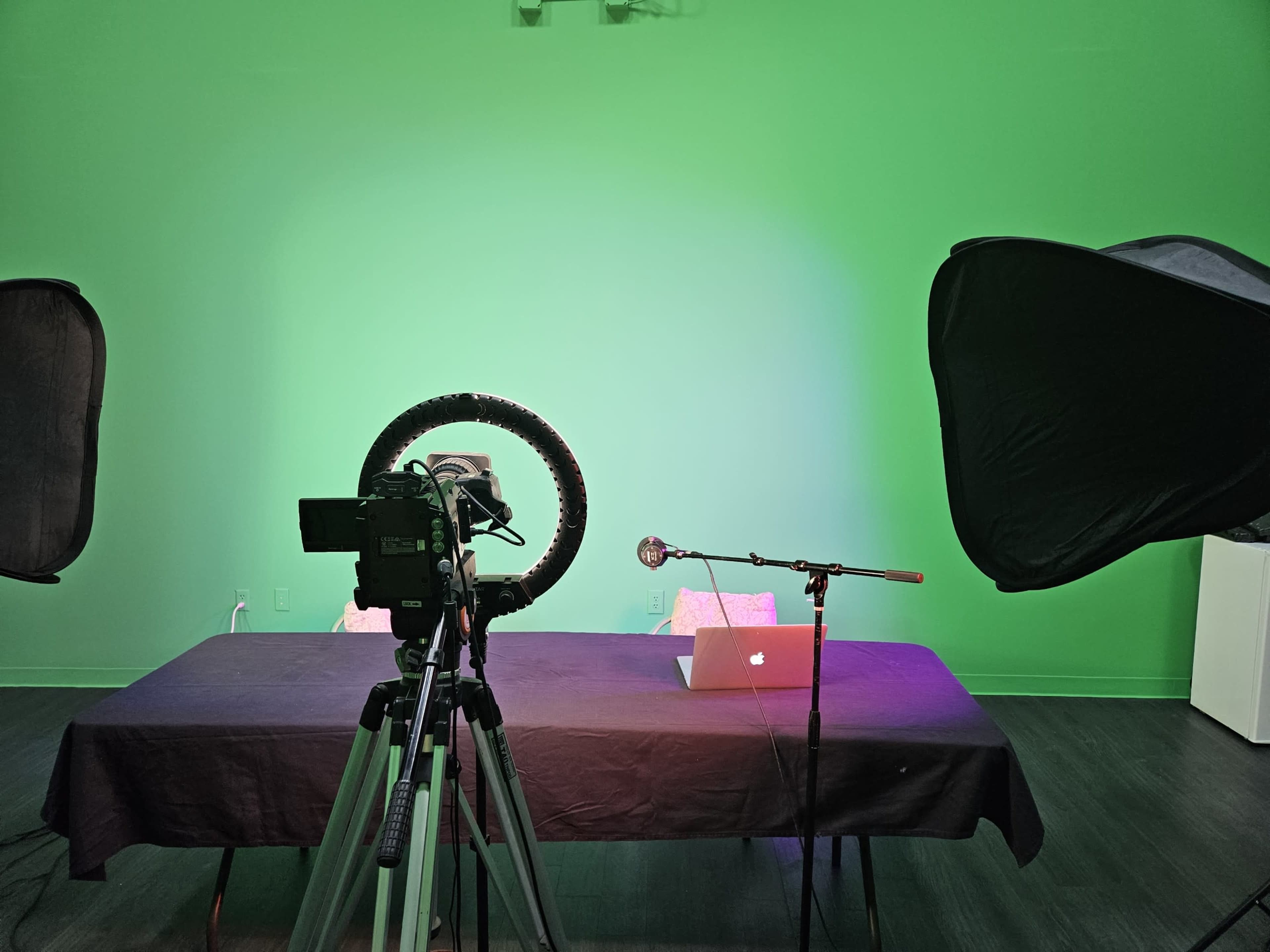 A video recording setup featuring a camera on a tripod, a microphone on a stand, two softbox lights, and a table with a laptop, all against a green backdrop.