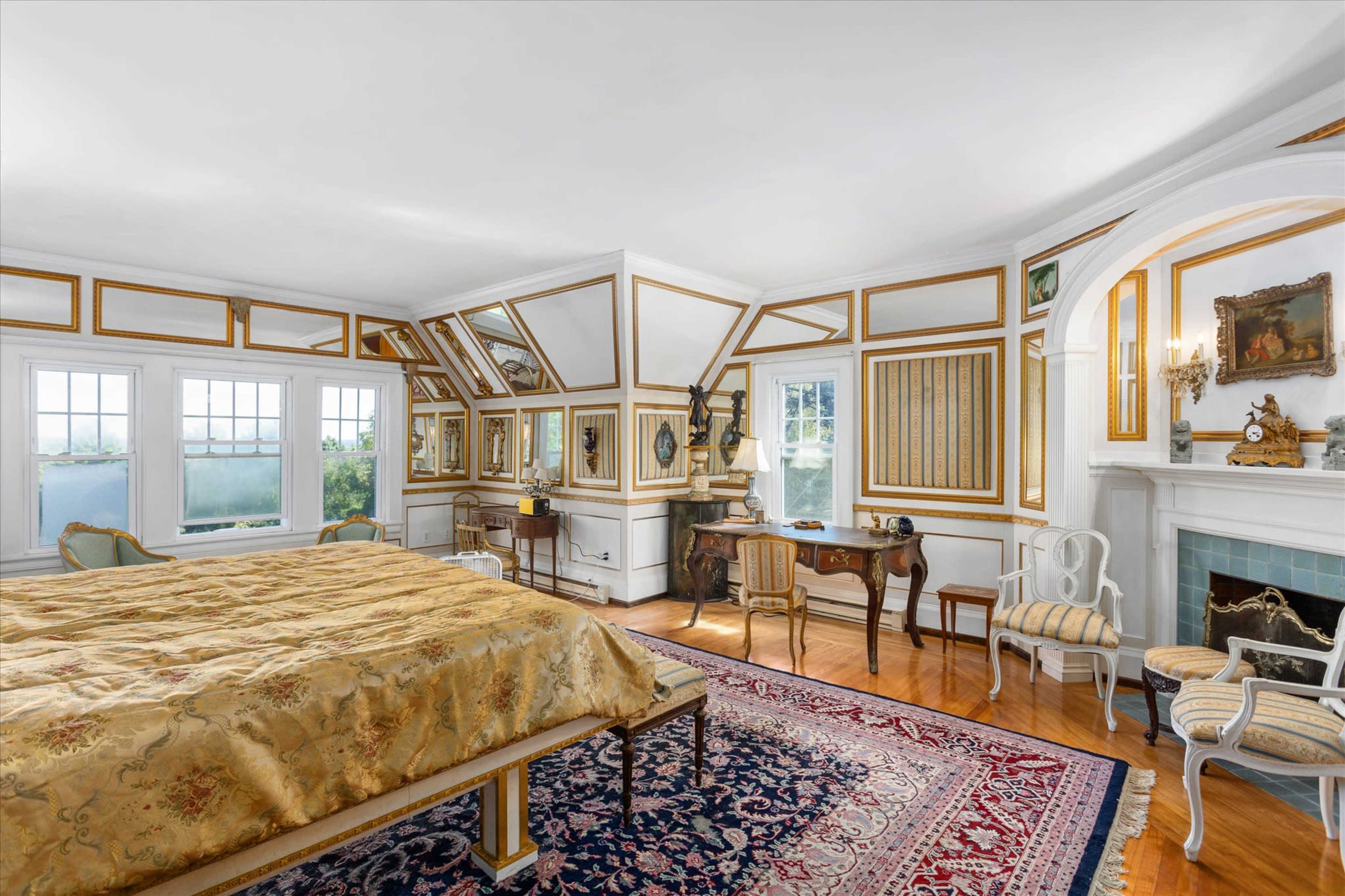 A spacious bedroom with a large bed, decorated walls featuring ornate trim, a vintage desk, and several stylized chairs arranged around a fireplace.