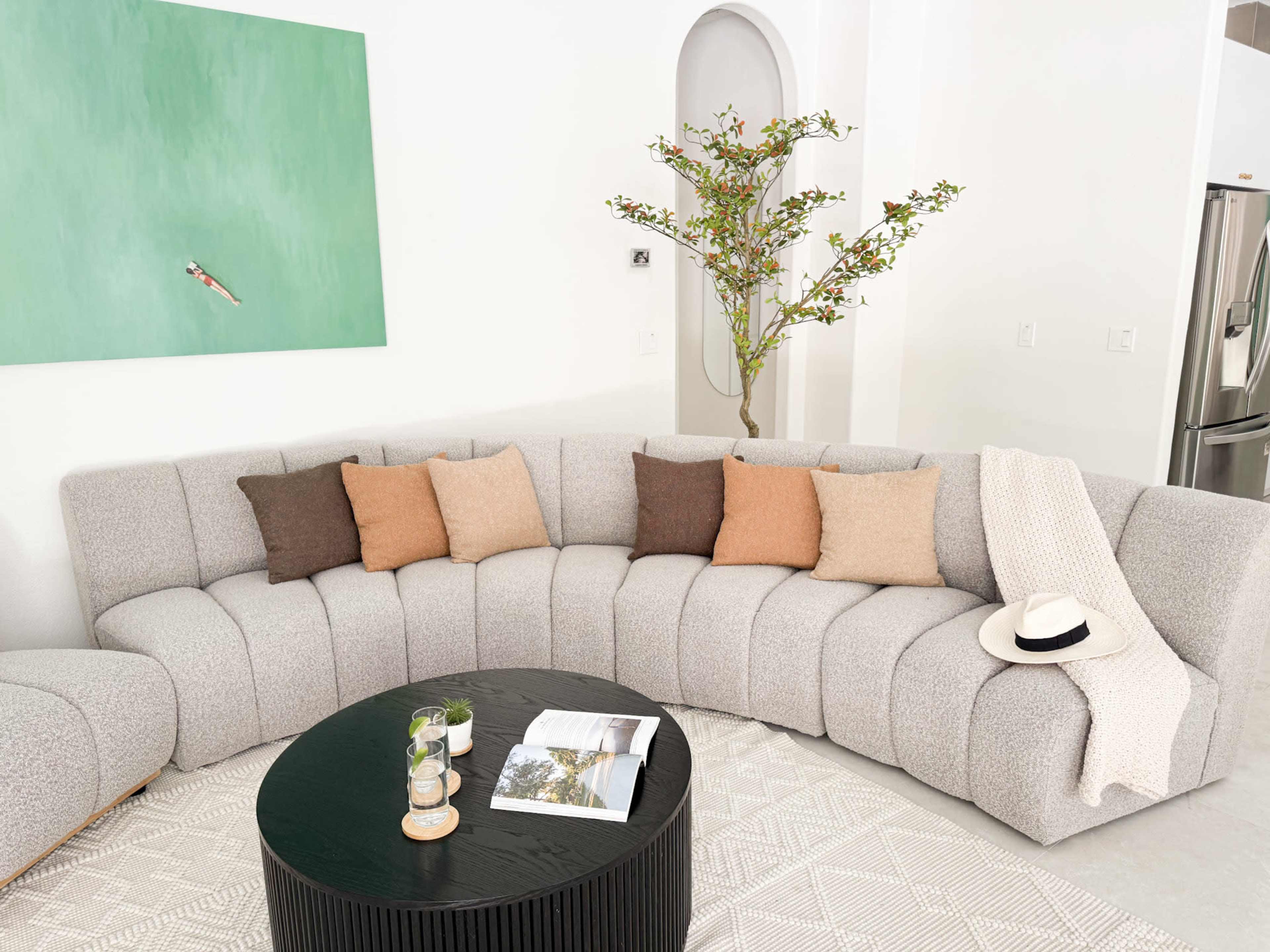 A curved sectional sofa with multiple throw pillows is arranged around a round black coffee table on a light-colored rug, with a green wall art piece and a potted plant in the background.