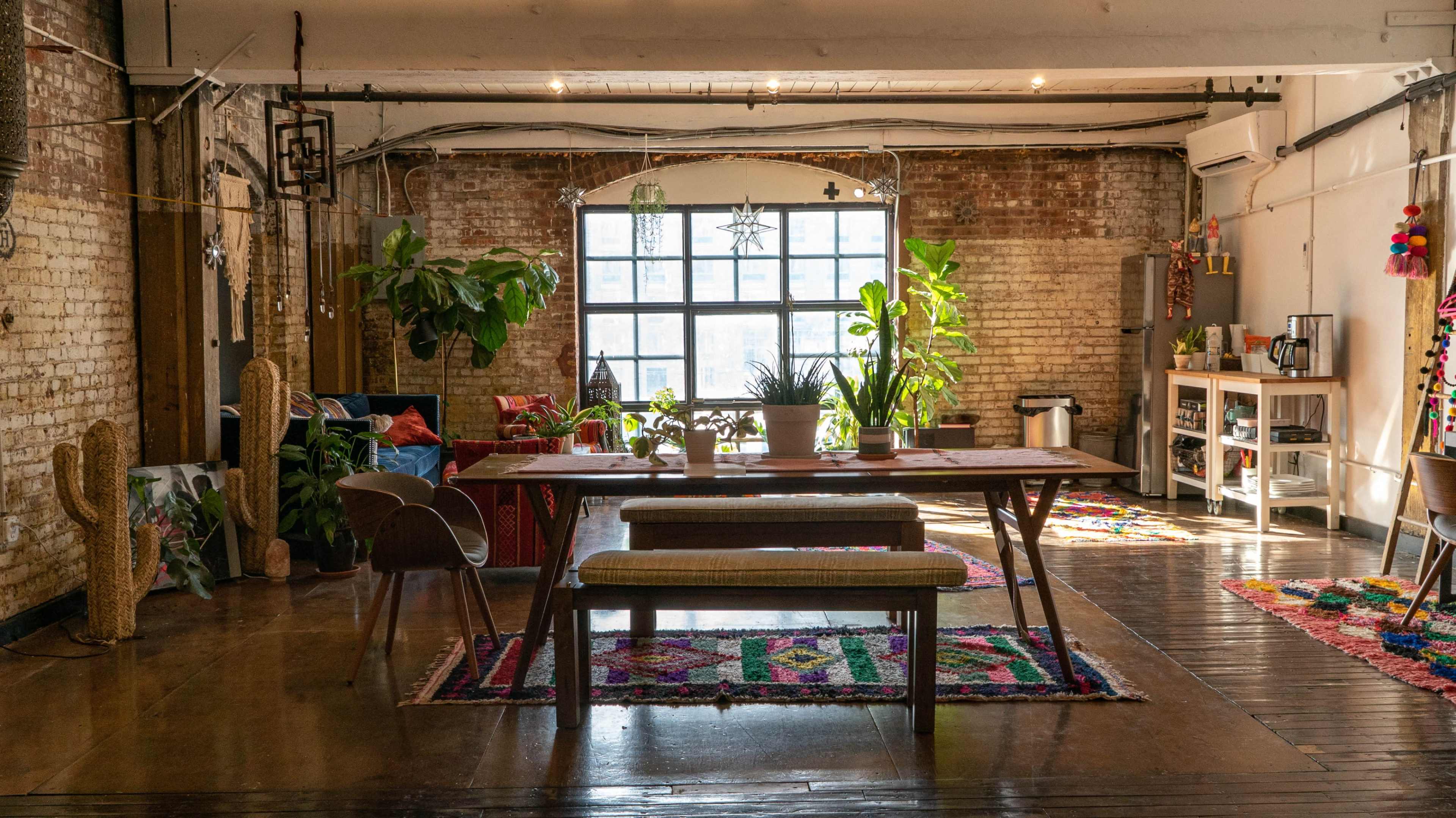 The image shows a spacious, well-lit room with a long wooden table and various plants, featuring a blend of modern and rustic decor.