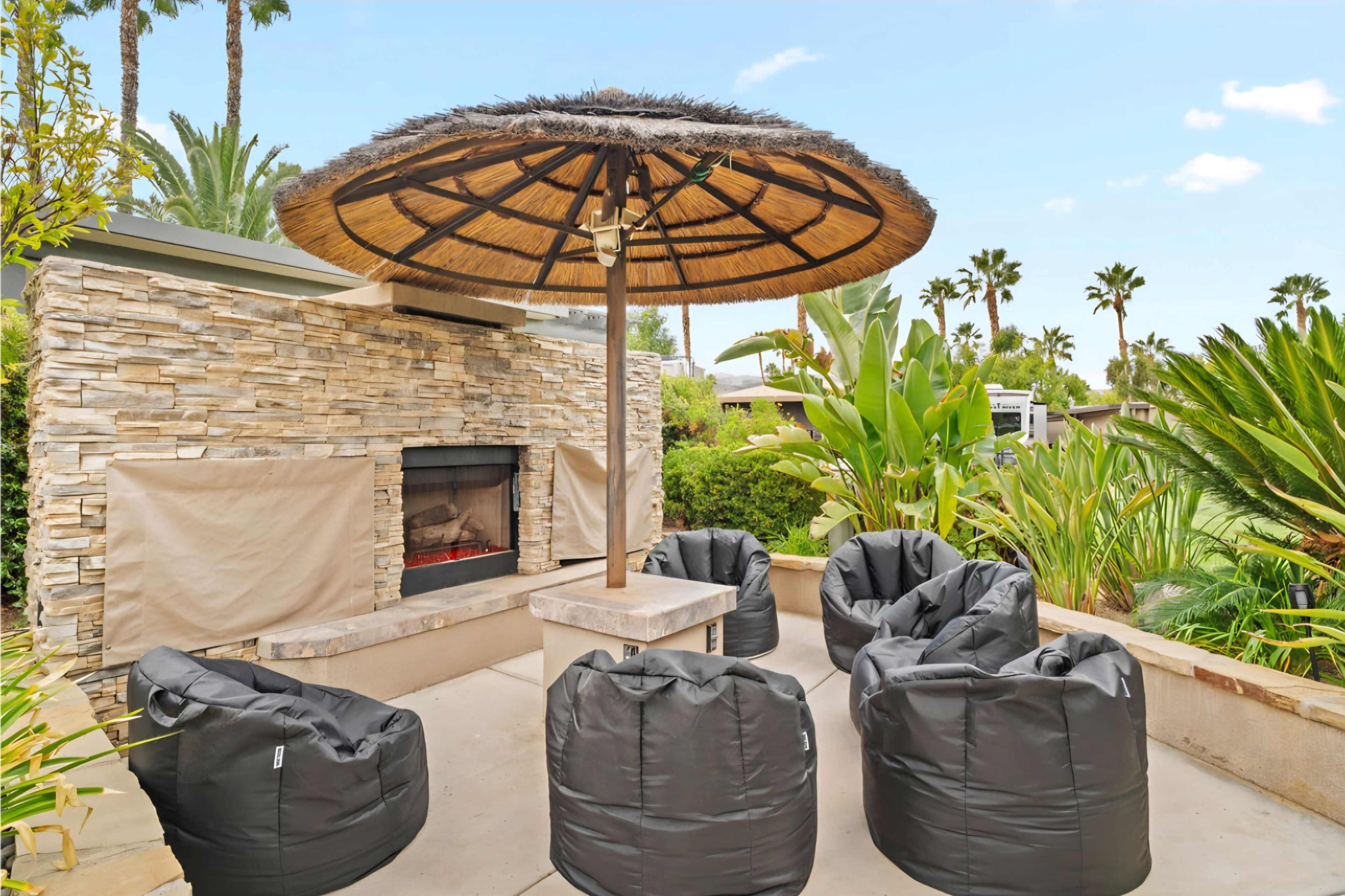The image shows a shaded outdoor seating area with a thatched umbrella, stone fireplace, and black bean bag chairs surrounded by tropical plants.