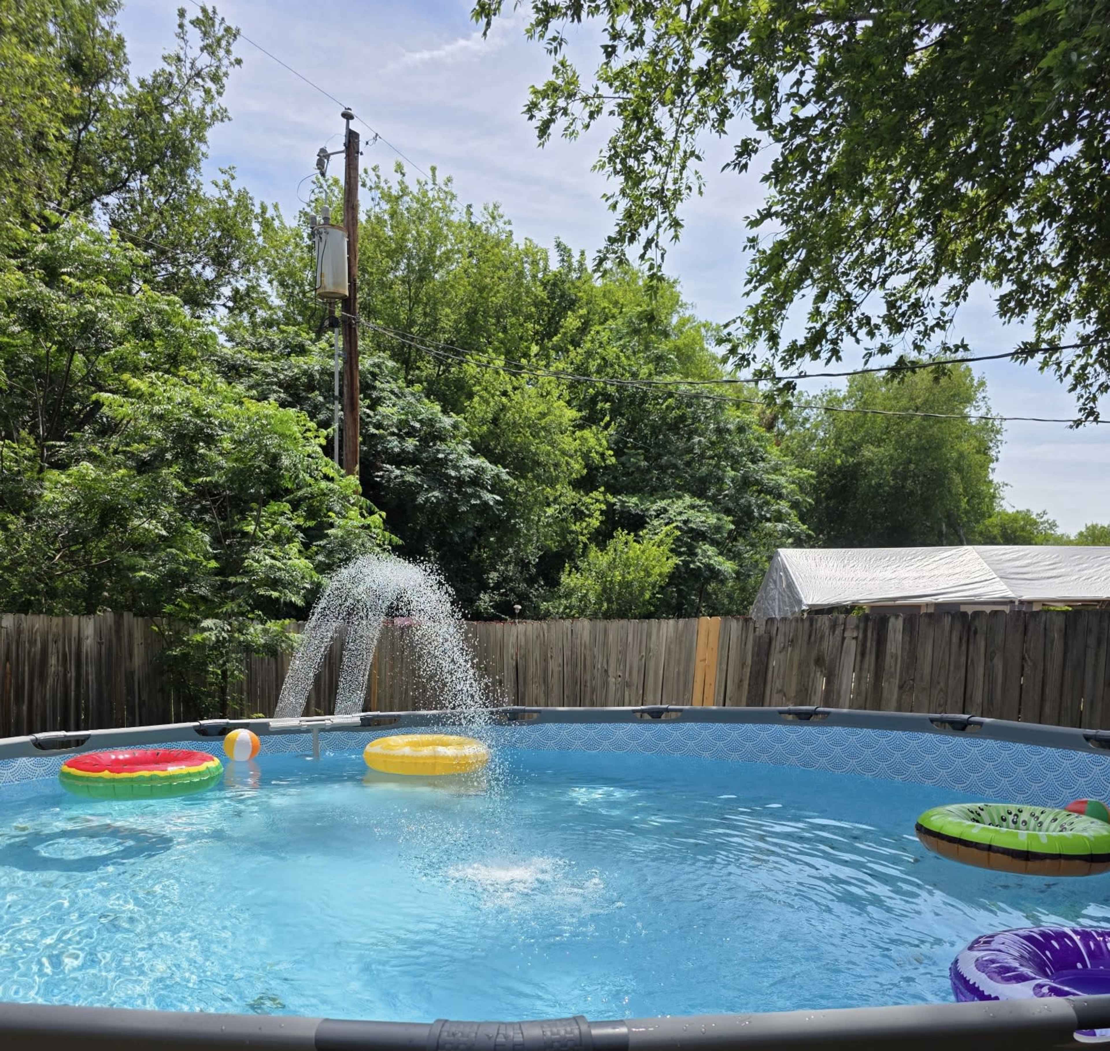 A round above-ground pool filled with blue water features floating pool toys and a fountain spray.
