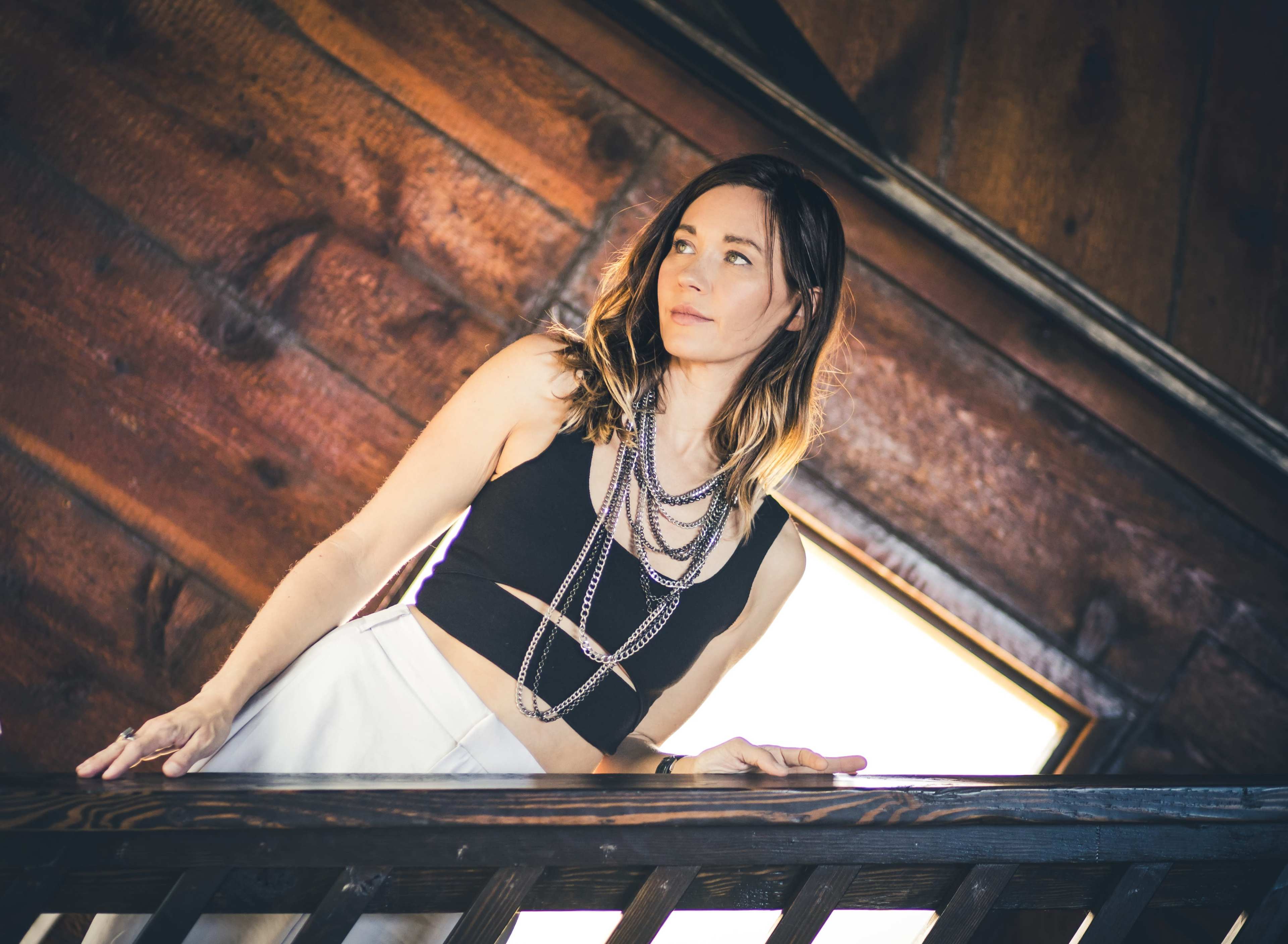 A woman wearing a crop top and layered necklaces stands by a wooden railing in an interior space with large windows.
