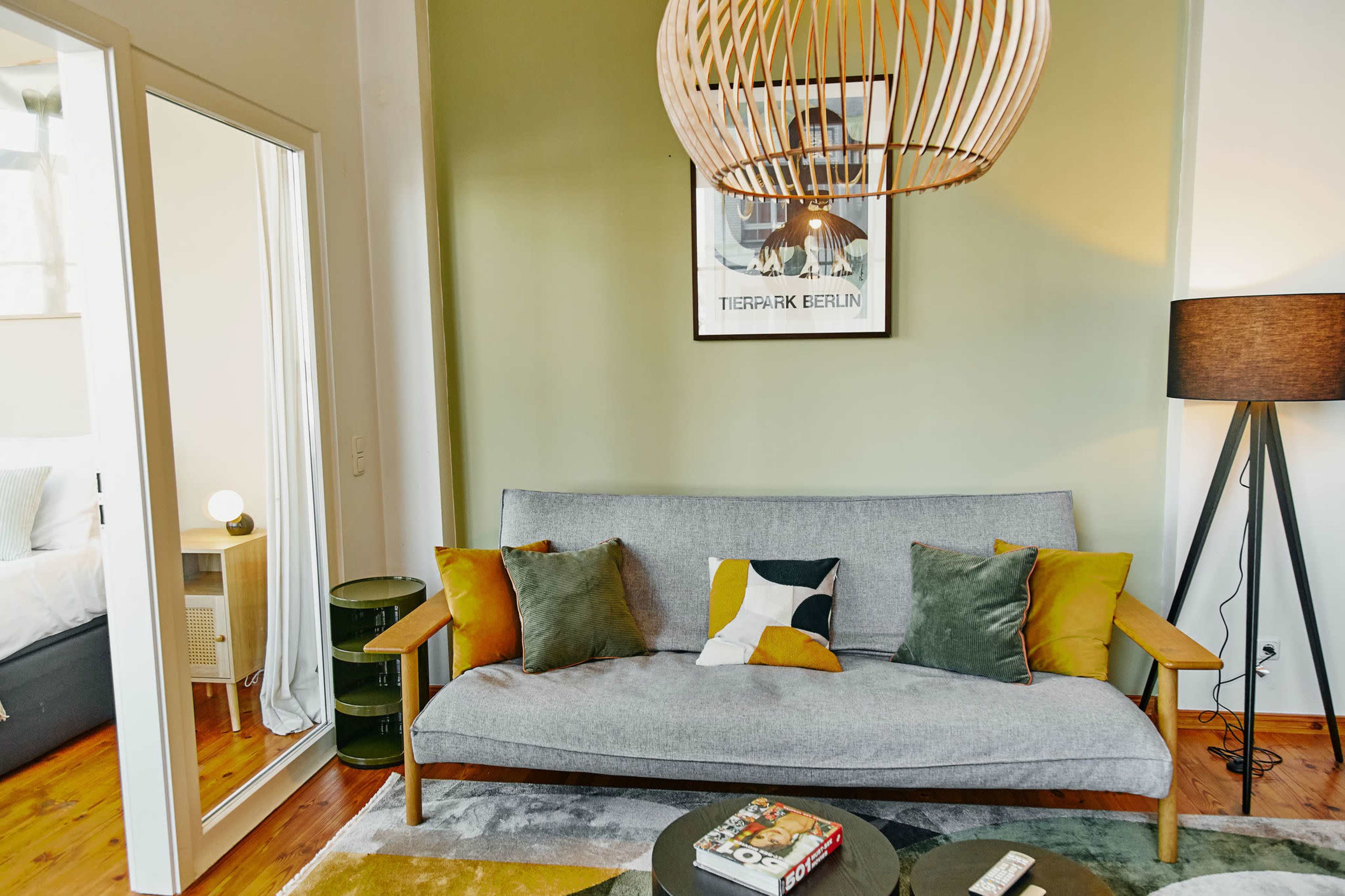 The image shows a modern living room featuring a gray sofa adorned with colorful pillows, a round coffee table, and a large pendant light overhead against a green accent wall.