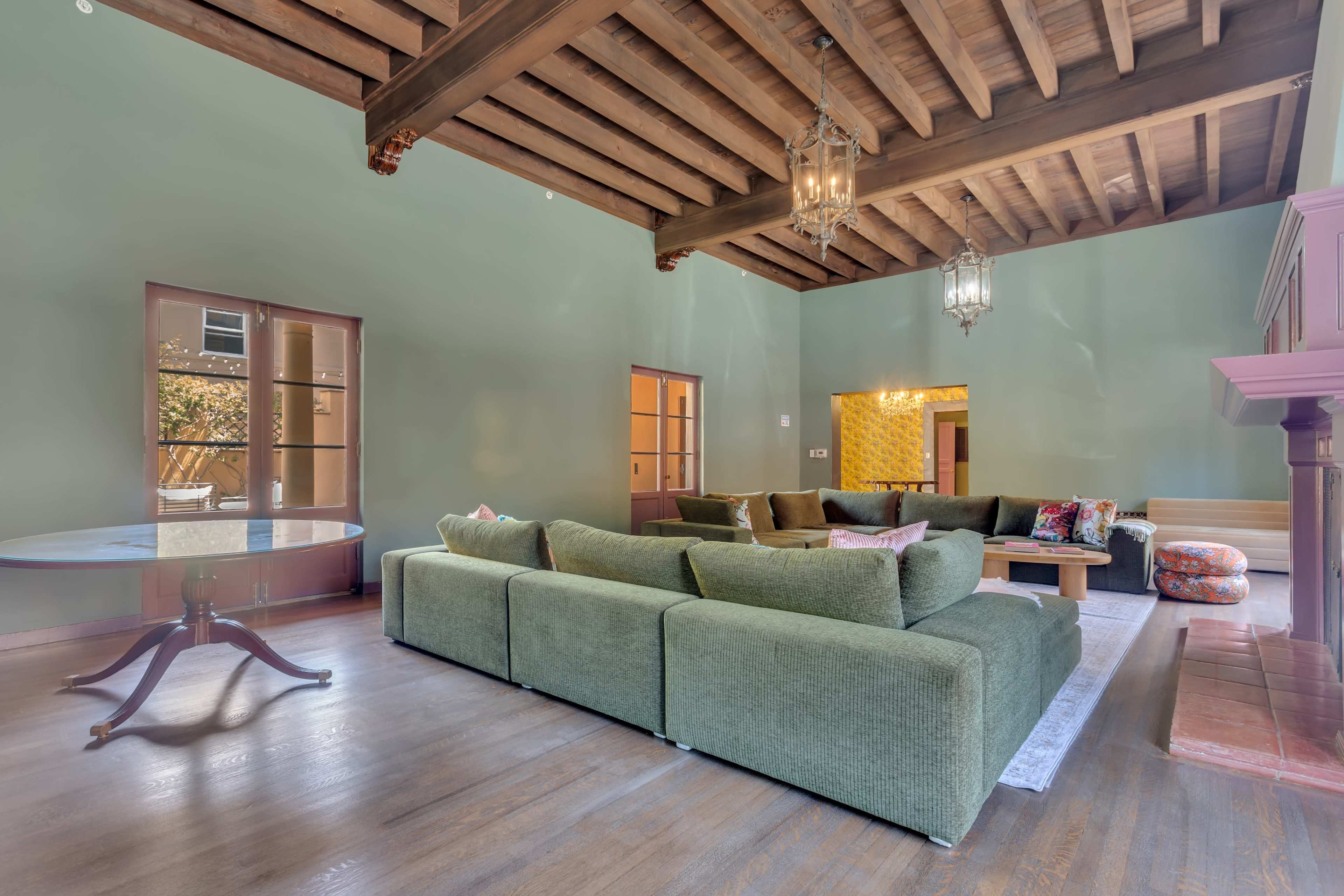 A spacious living room features a large green sectional sofa, wooden ceiling beams, and multiple windows with natural light.