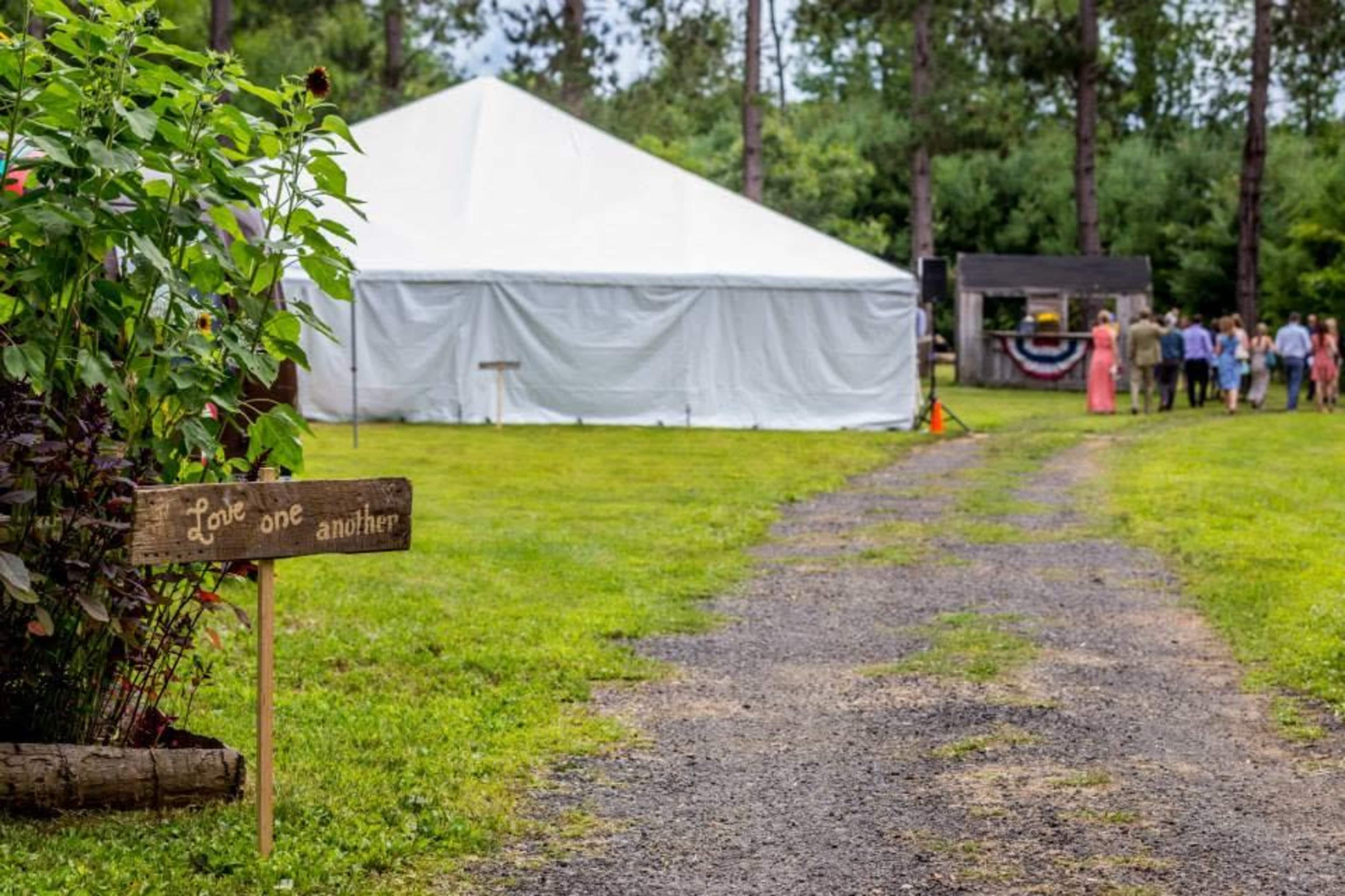 A gravel path leads to a large white tent set in a grassy area surrounded by trees, with a sign that reads "Love one another."
