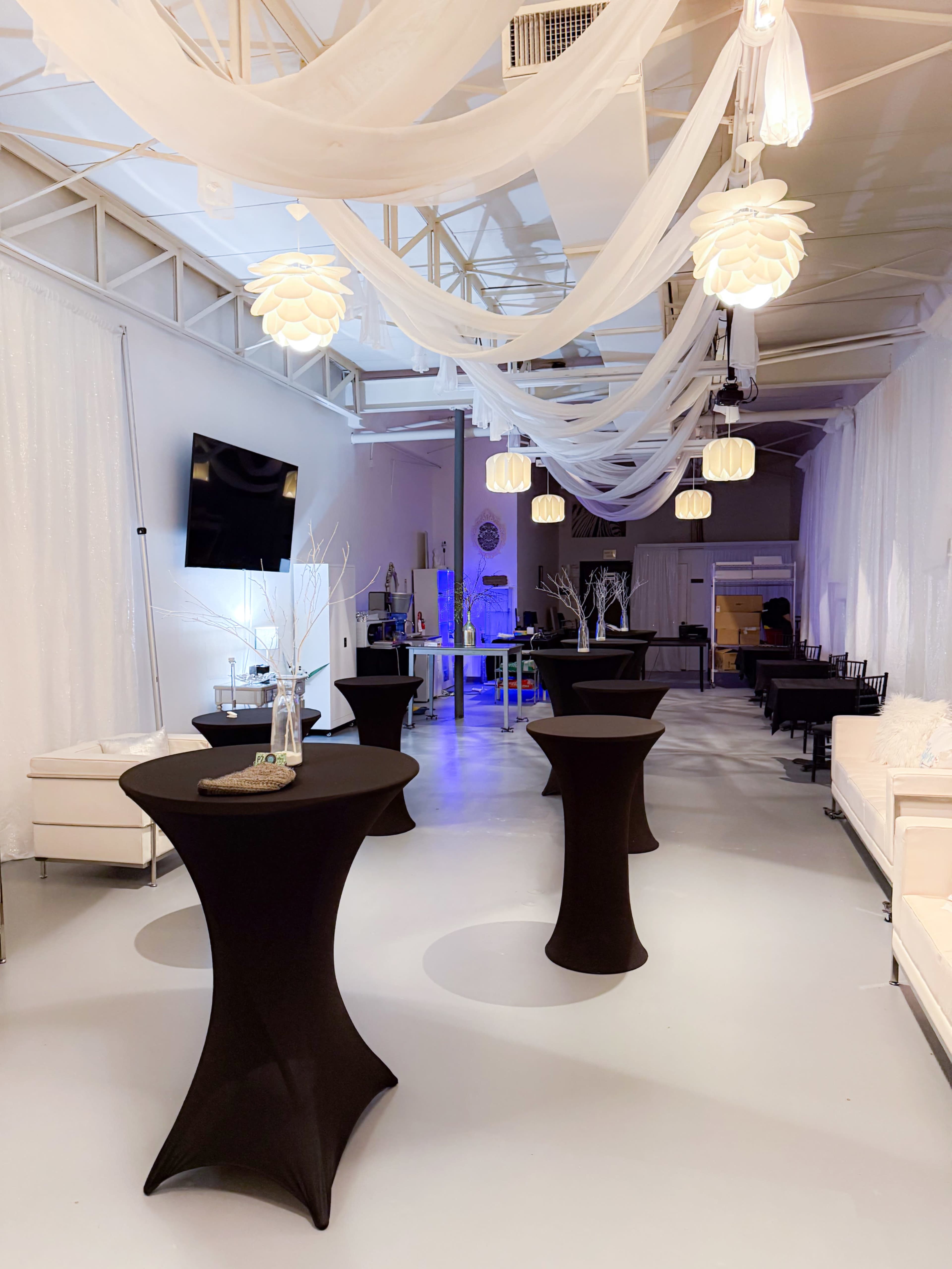 The image shows a spacious indoor event venue decorated with sheer drapery, pendant lights, and black cocktail tables arranged throughout the room.