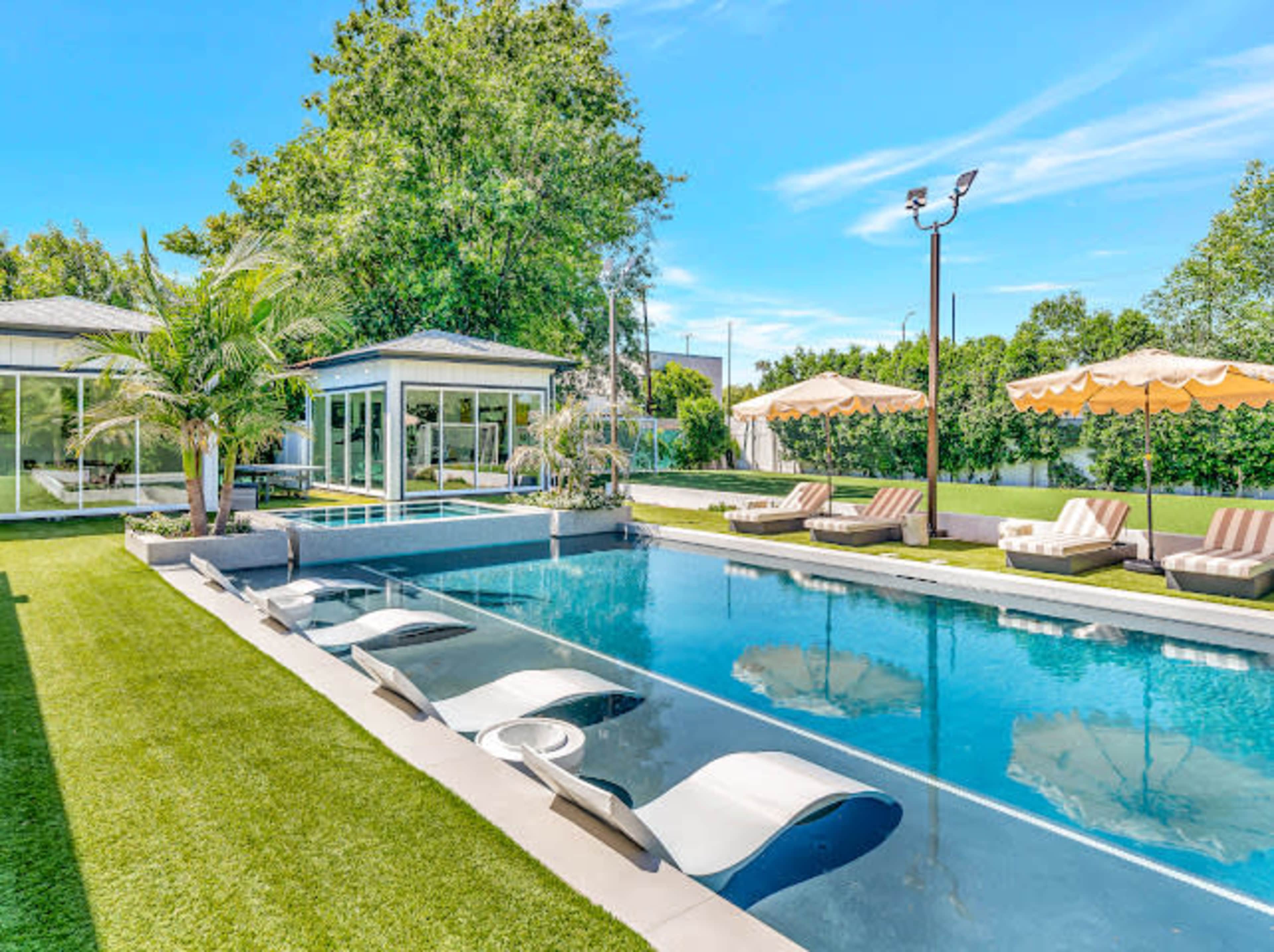 The image shows a modern outdoor pool area featuring lounge chairs, palm trees, and a glass enclosure with umbrellas in a lush green setting.