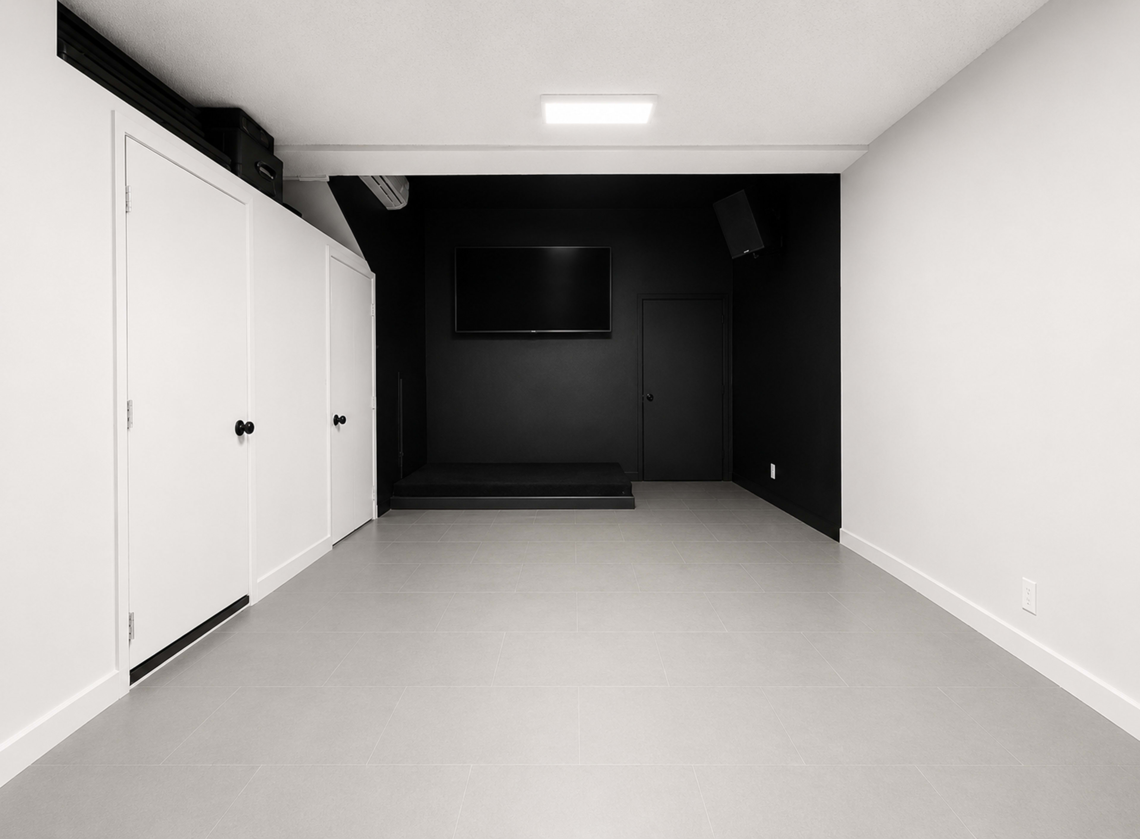 The image shows a minimalist room with a dark wall, a mounted television, and a platform, alongside white doors and light gray tile flooring.