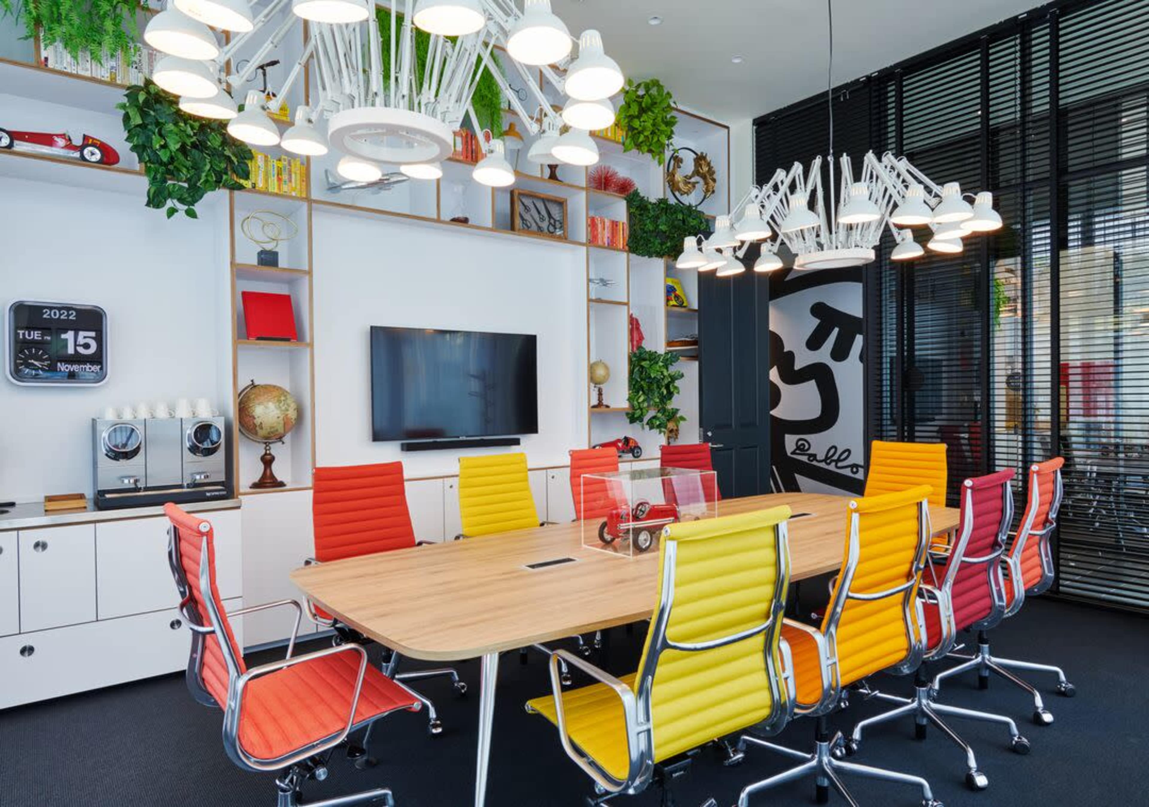 A bright conference room features a large wooden table surrounded by colorful chairs, with a wall-mounted TV and decorative plants.