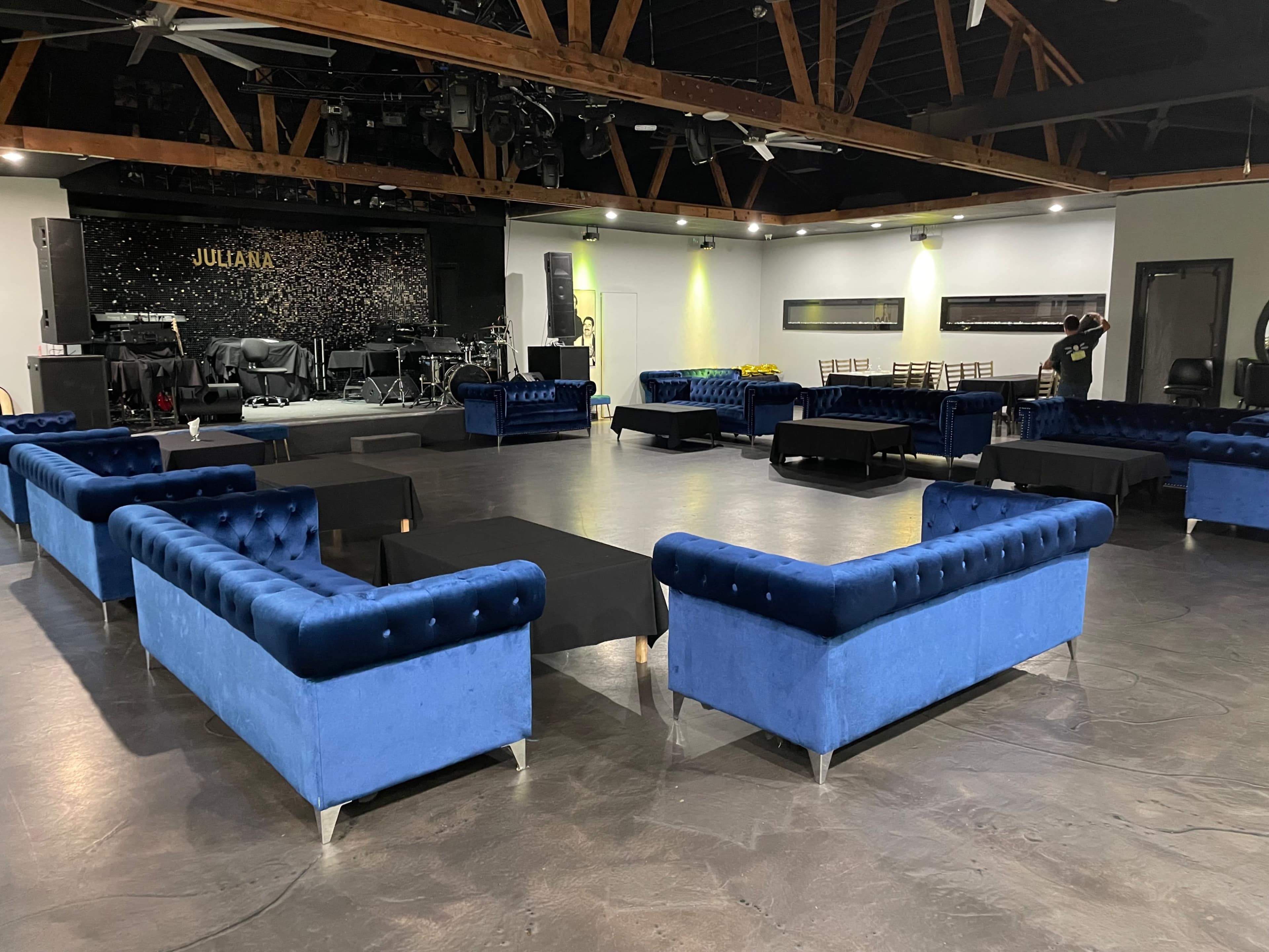 A spacious venue with blue velvet couches arranged around black coffee tables, set up for an event with a stage in the background.
