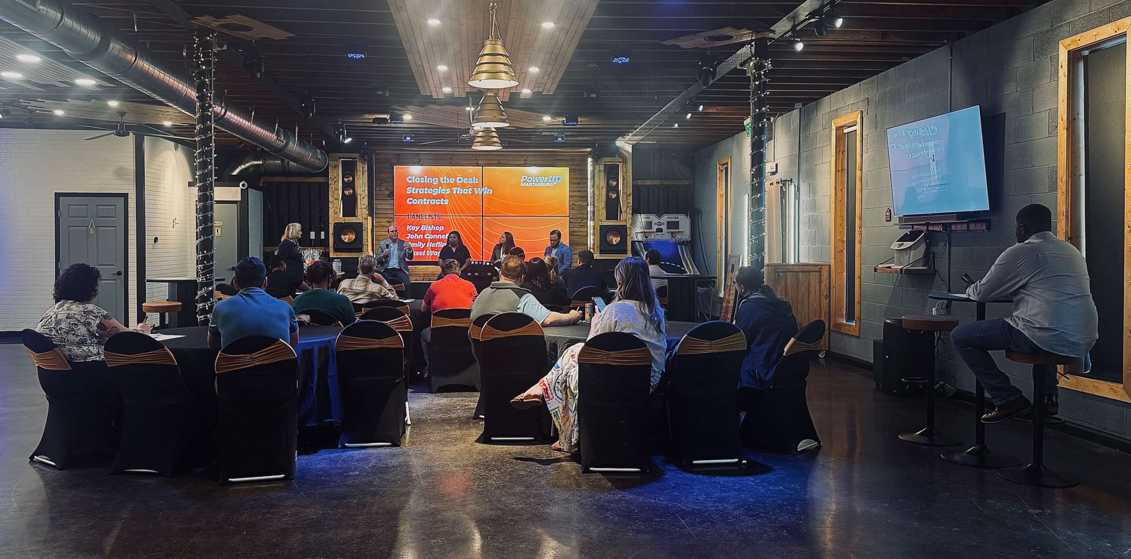 A group of people is seated in a conference room, attentively facing a large presentation screen displaying information about closing deals and strategies for financial growth.