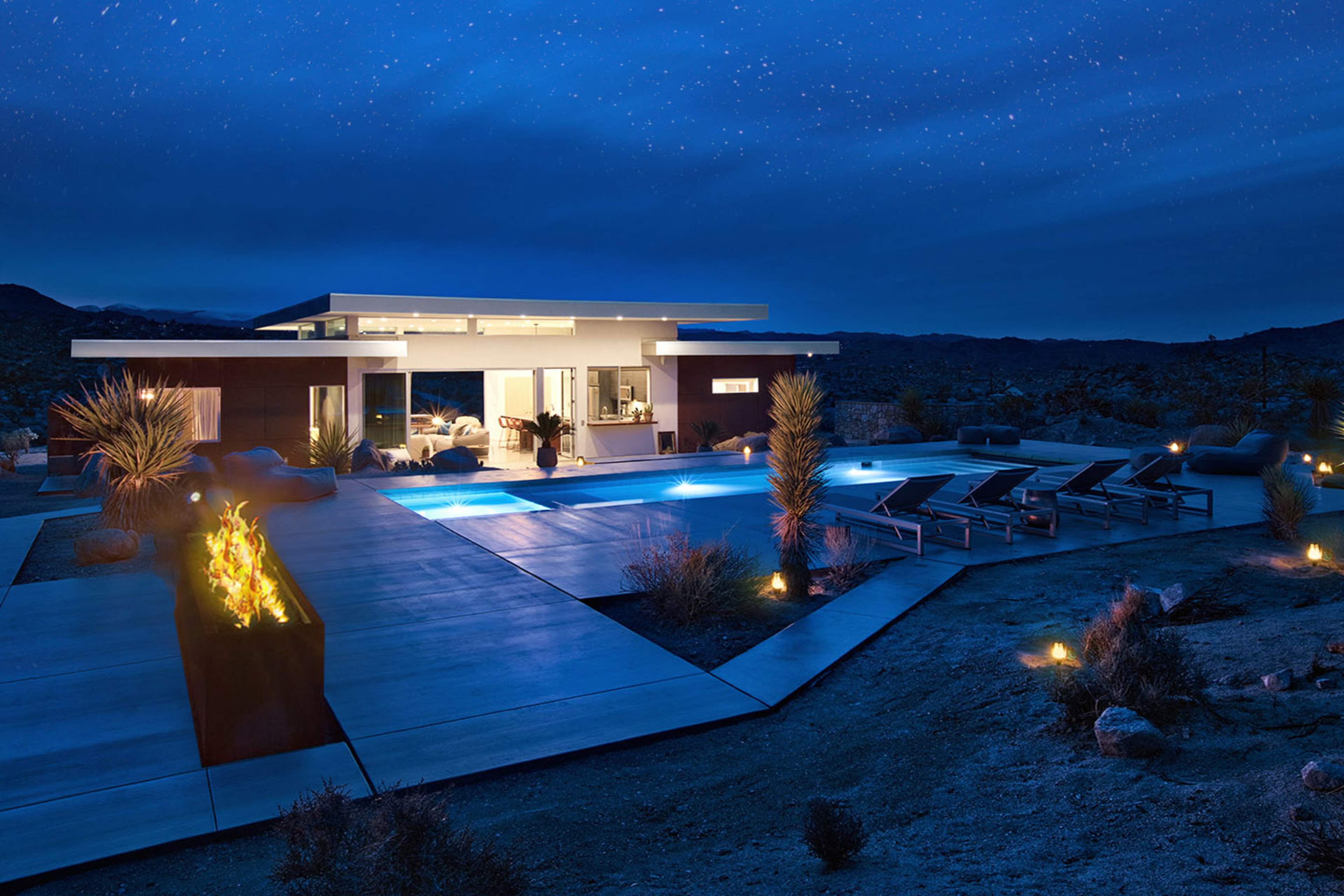 A modern home with a pool and outdoor seating is illuminated under a starry night sky in a desert landscape.