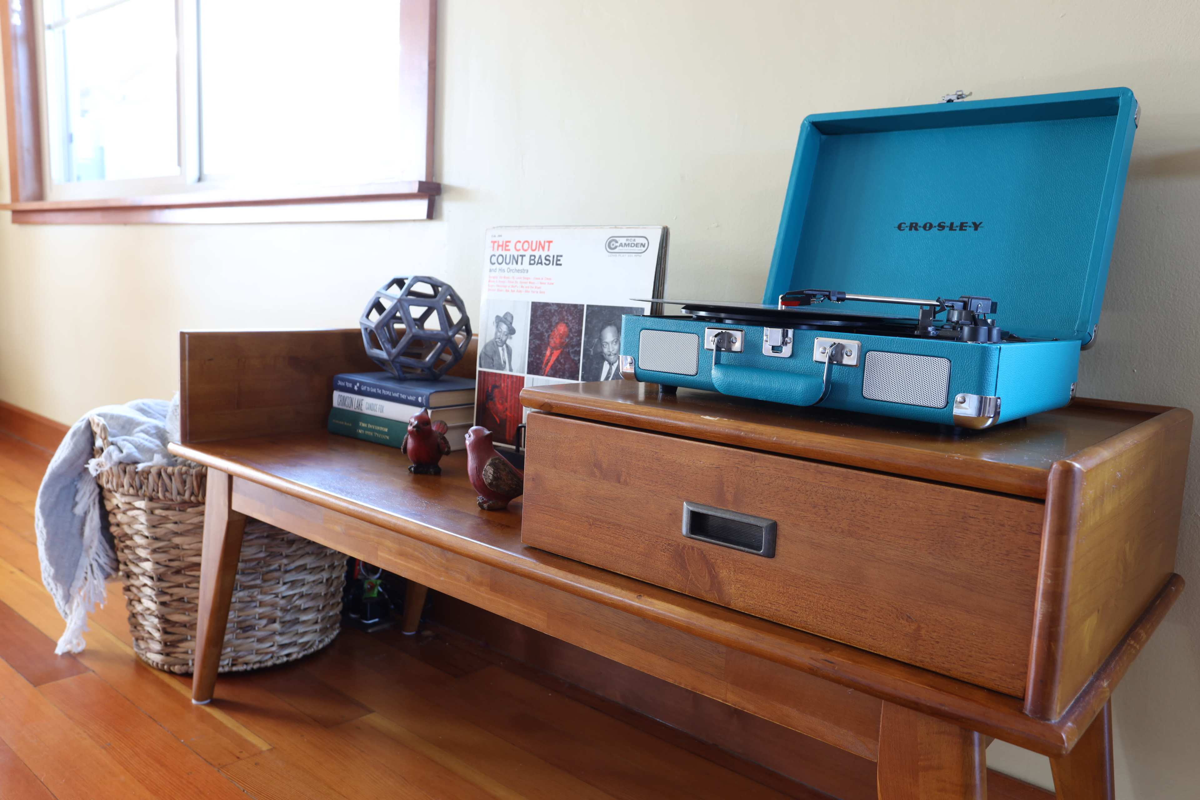 A wooden bench with a record player, decorative items, and a basket sits beside a window.