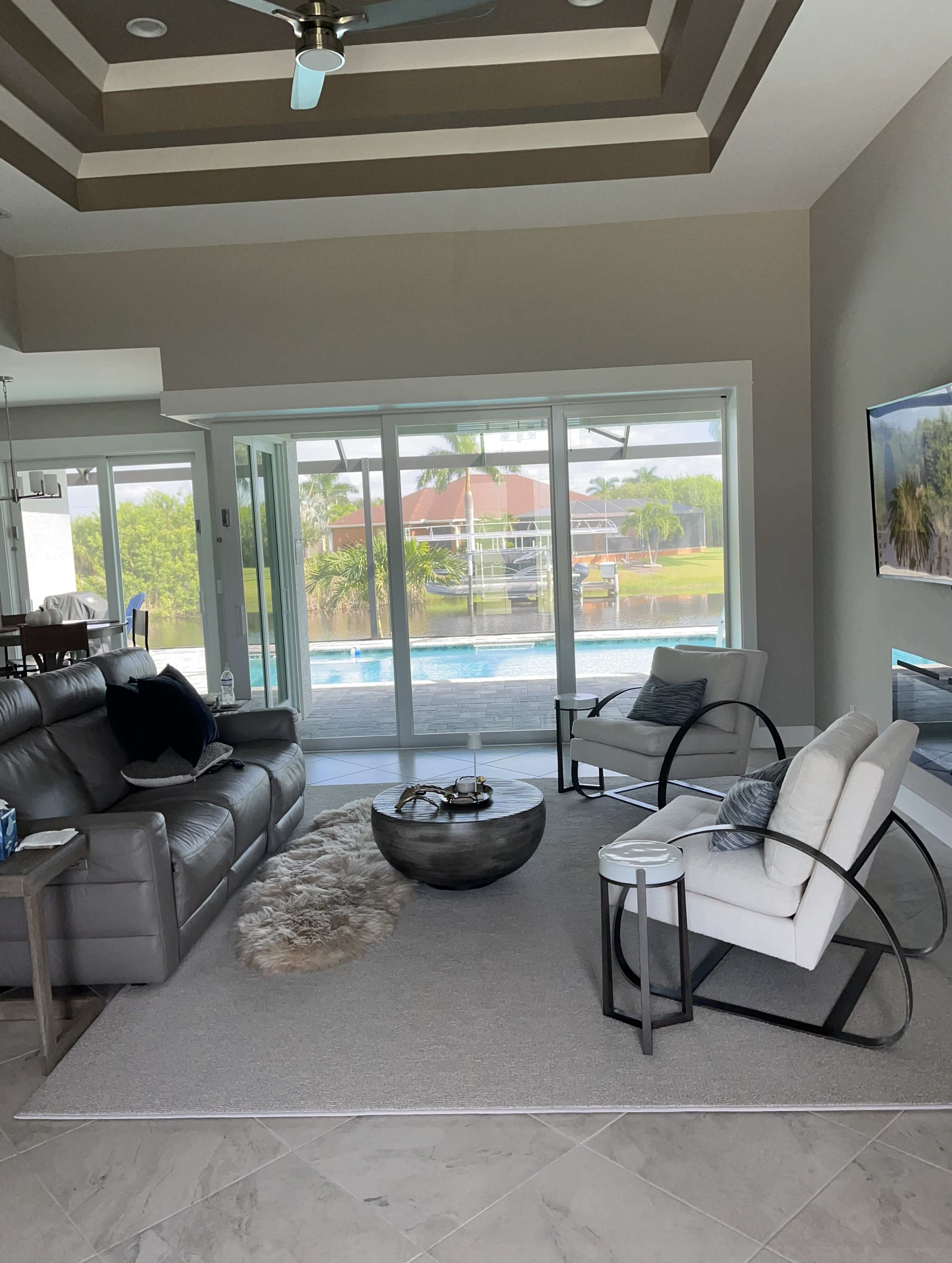 The living room features a gray sofa and two white armchairs around a round coffee table, with large windows that overlook a pool and patio area.