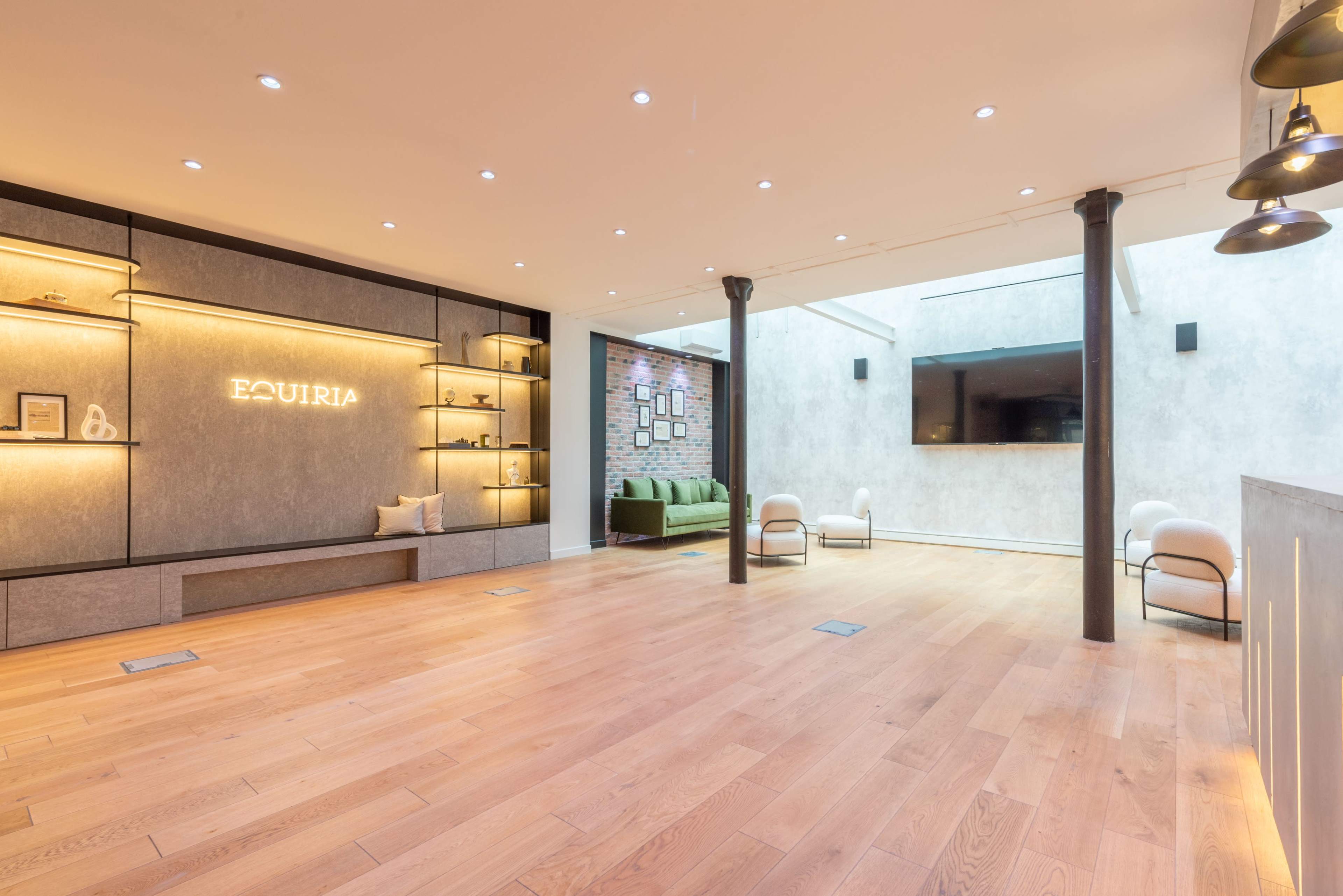 The image shows a modern, minimalistic interior space with wooden floors, wall shelves illuminated with warm lighting, and a green couch along the back wall.