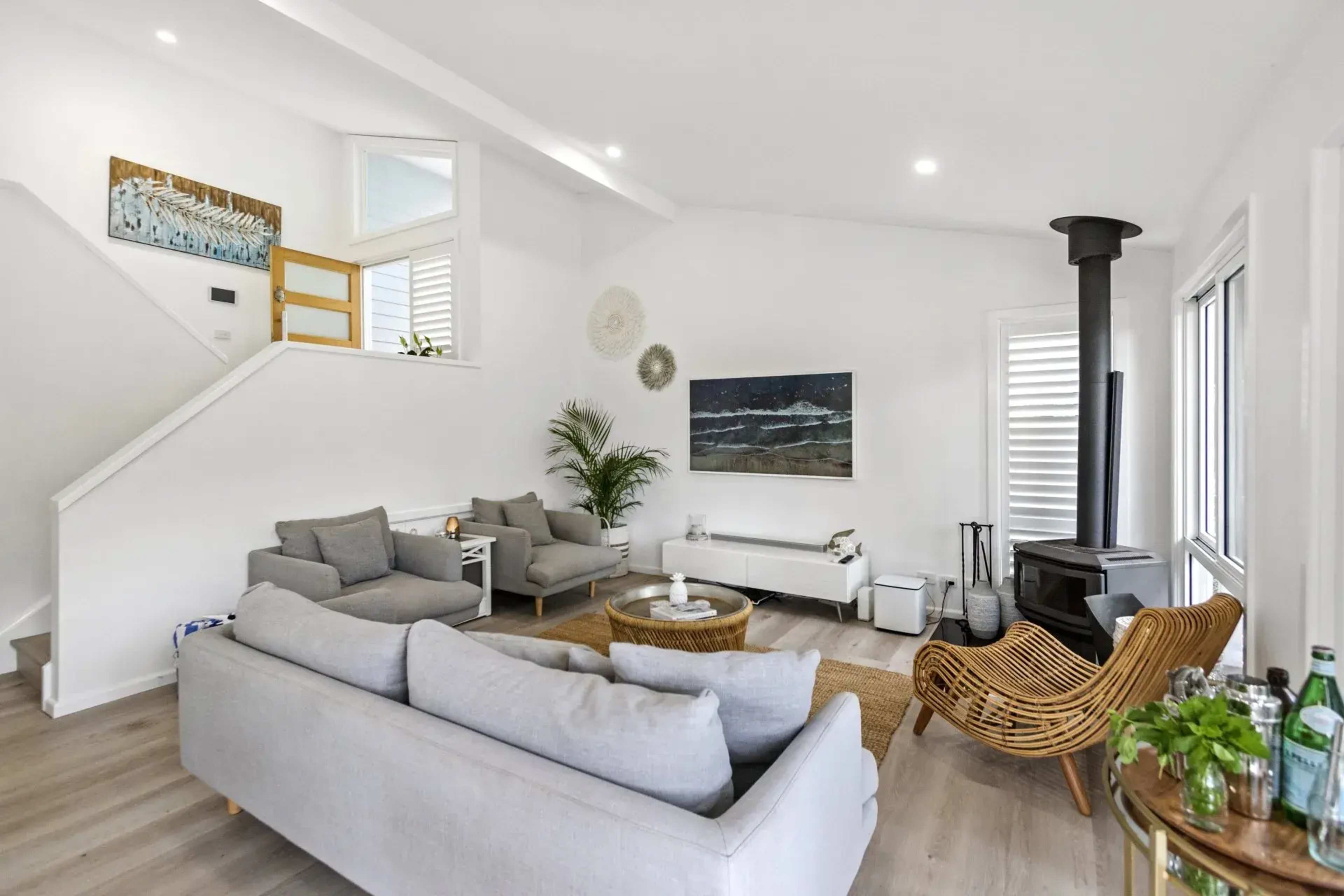 A contemporary living room with a gray sofa, a rattan chair, a coffee table, and a black wood-burning stove, all set in a bright space with large windows.