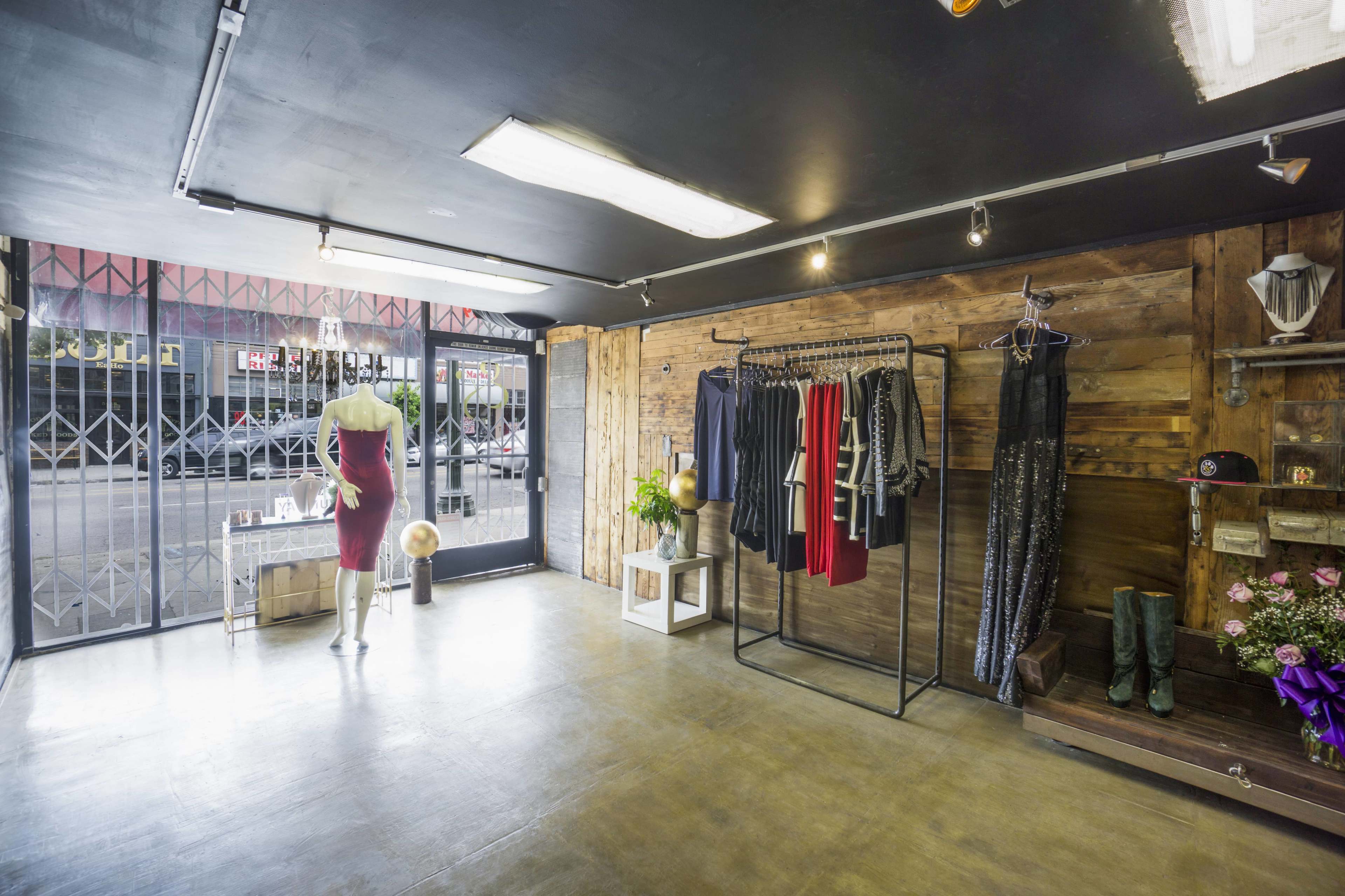 A clothing store with a mannequin displaying a red dress, clothing racks, and wooden walls, while a gated entrance opens to the street outside.