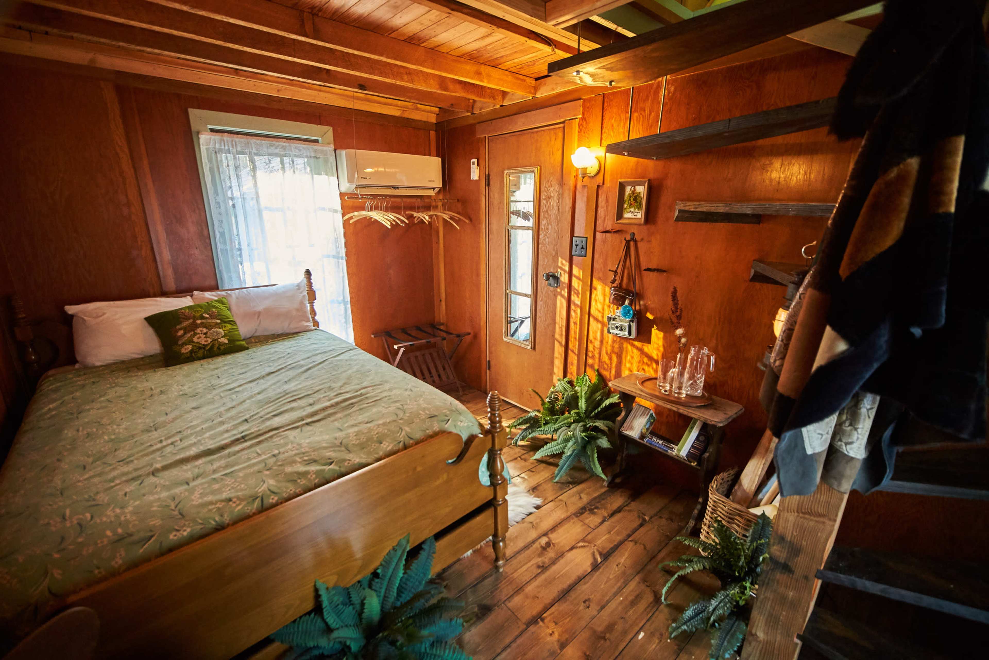 A cozy bedroom with a wooden bed, a small table, and plants, illuminated by warm light from a nearby window and a bulb.