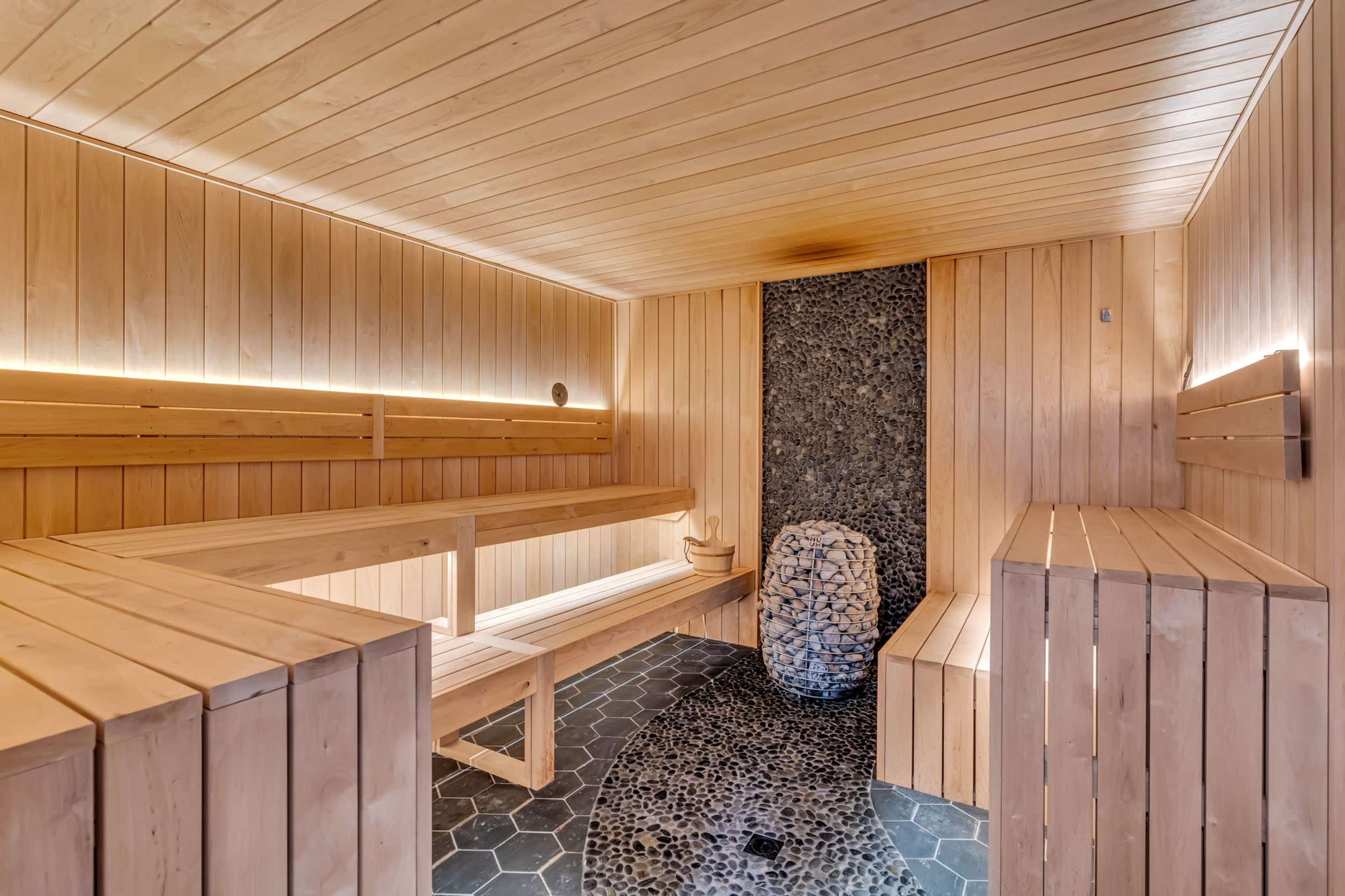 The image shows a wooden sauna with benches, a stone centerpiece, and illuminated wall features.