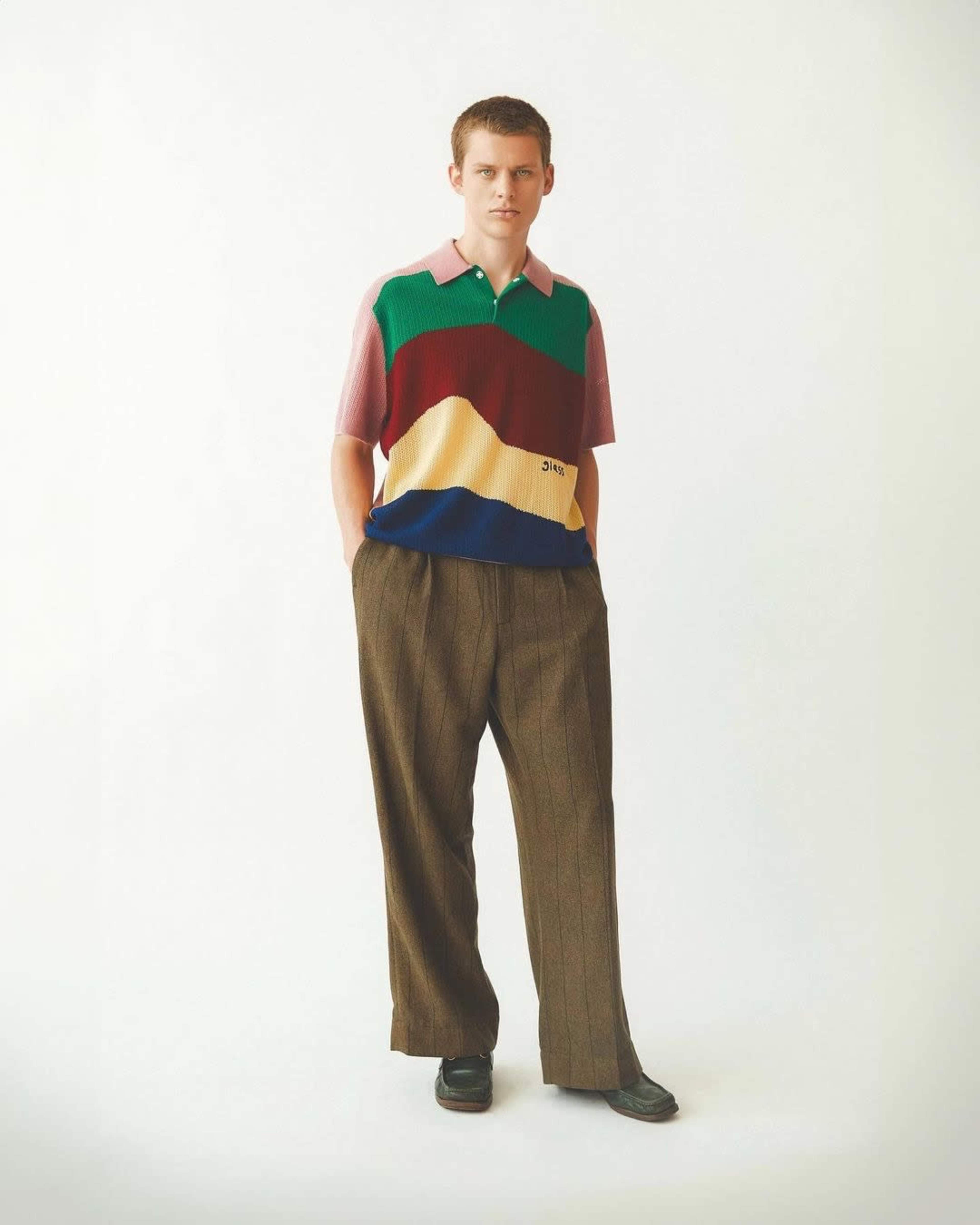 A young man stands wearing a multicolored, striped polo shirt and brown, pinstriped trousers.