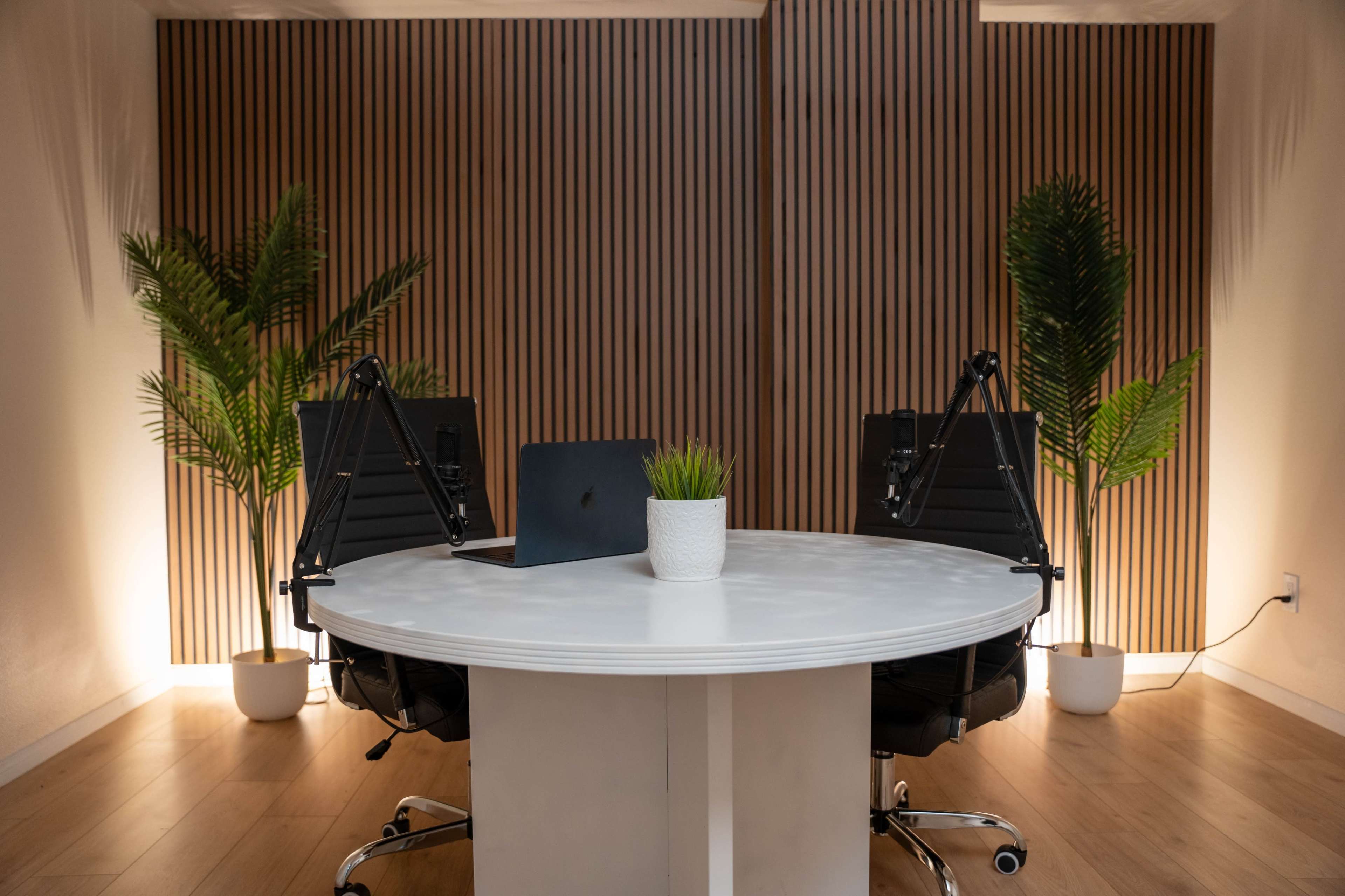 A modern workspace features a round white table with two black chairs, a laptop, and potted plants against a backdrop of wooden paneling and soft lighting.