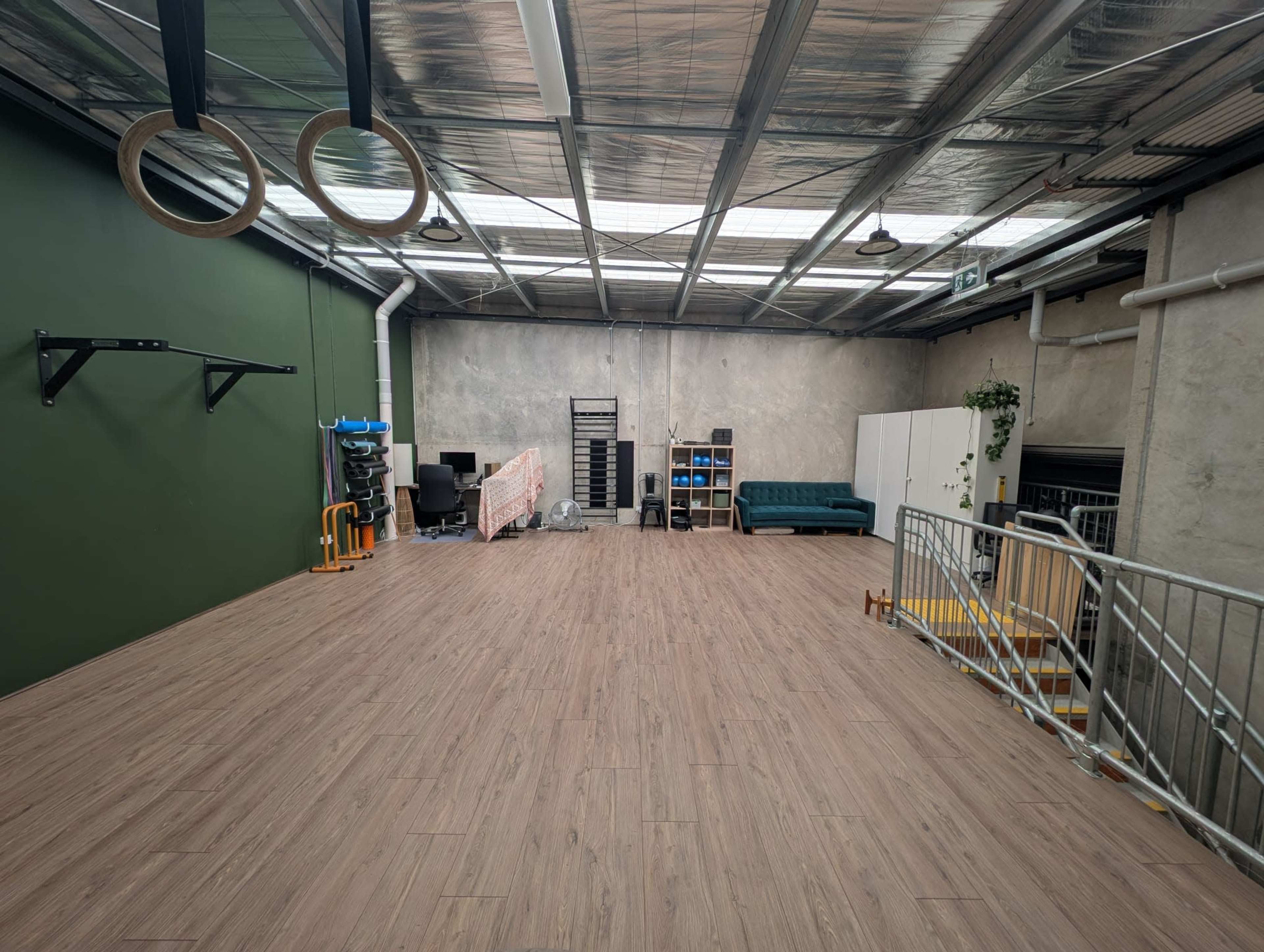 The image shows a spacious, industrial-style room with a polished wooden floor, green accent wall, gym equipment on one side, and various furniture, including a couch and office chairs, along the back wall.