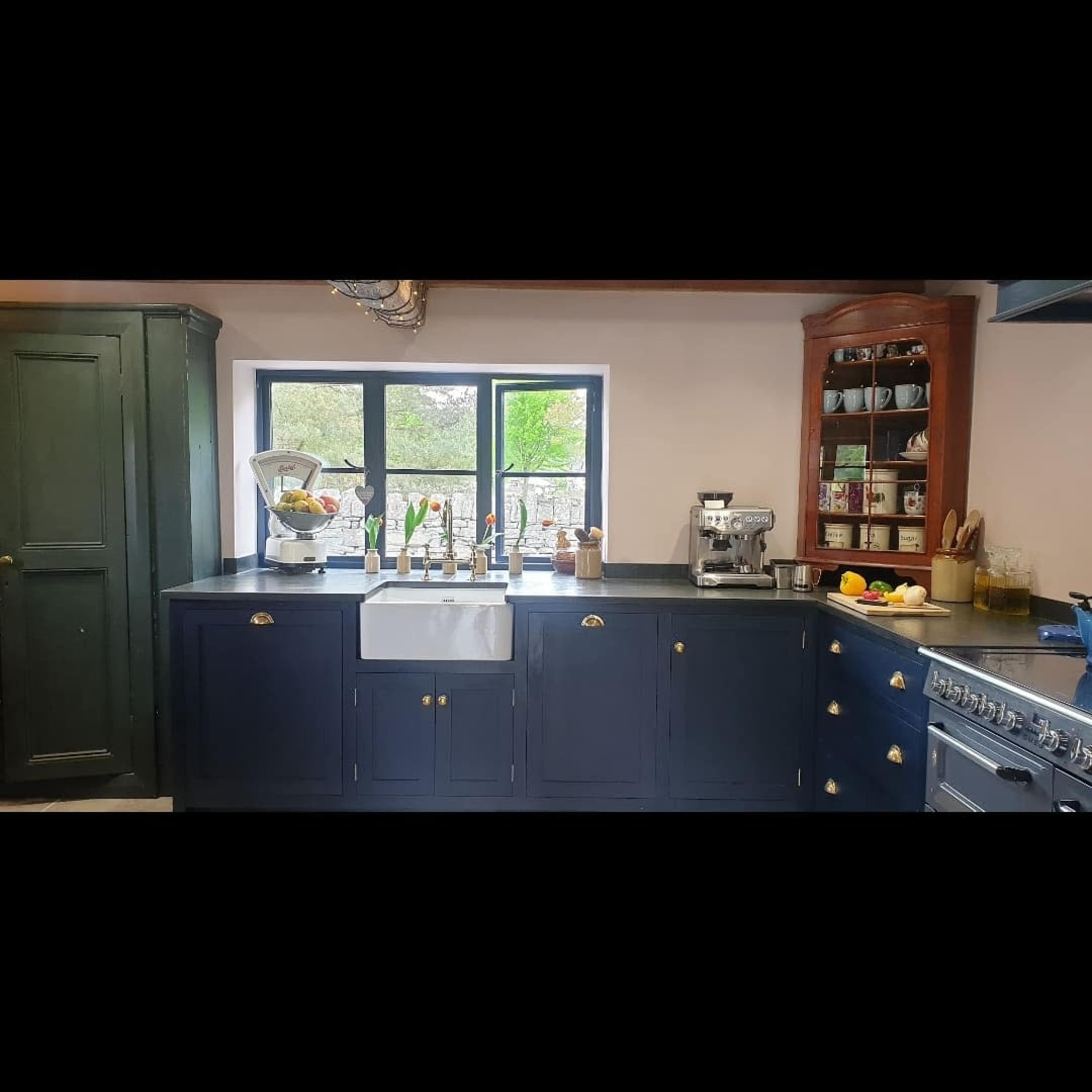 A kitchen with a navy blue countertop and cabinetry, featuring a farmhouse sink, a coffee machine, and a window with a view of greenery.