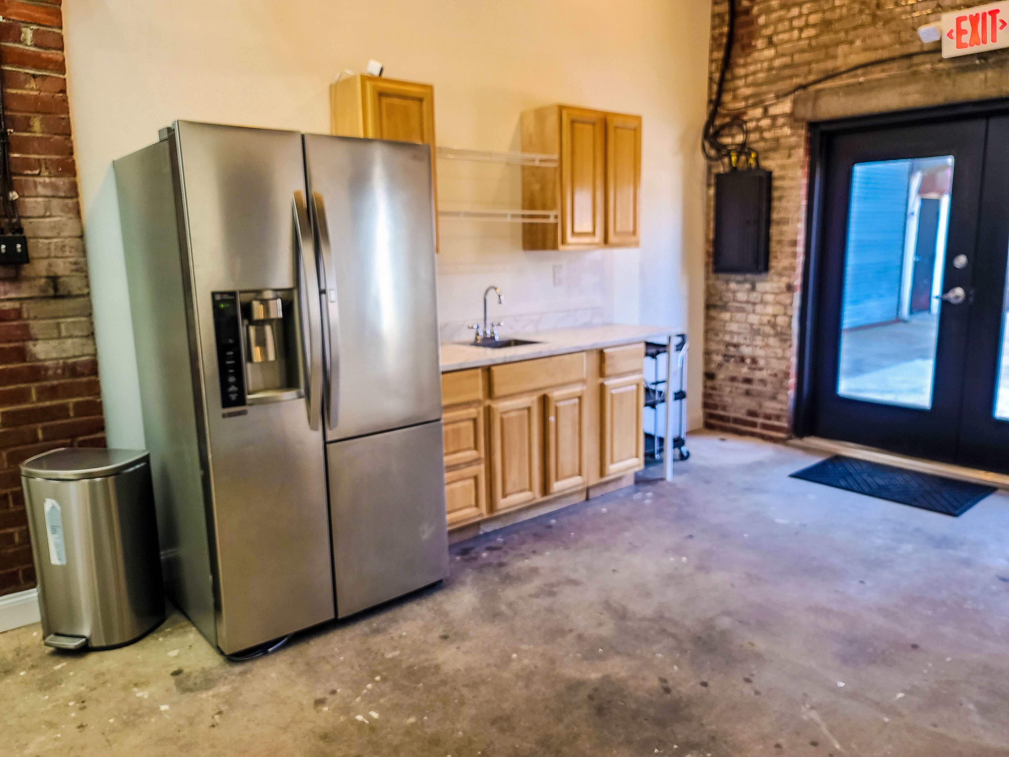A modern kitchen area featuring a stainless steel refrigerator, wooden cabinets, a sink, and a door leading outside in an industrial-style space.