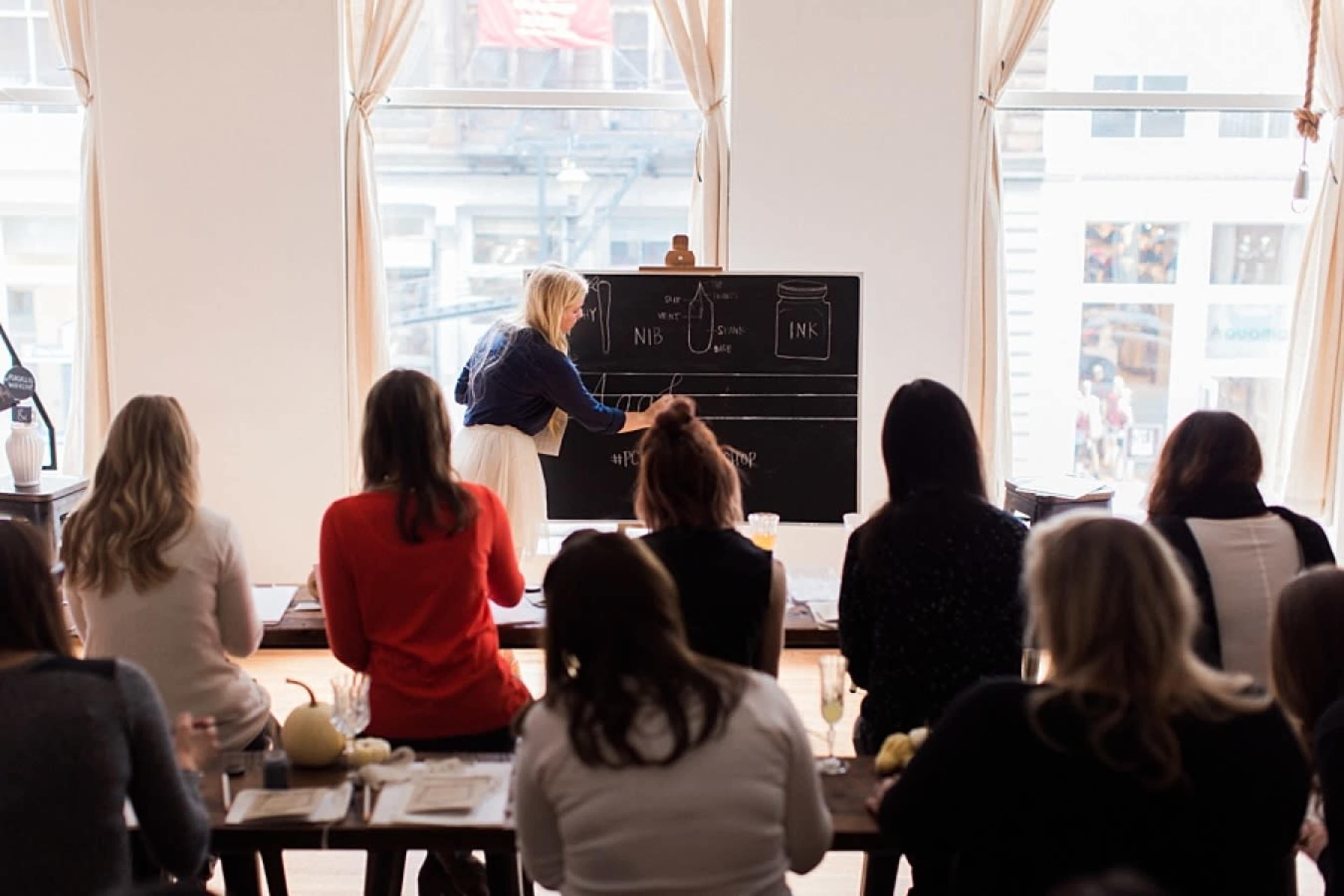 A woman writes on a large blackboard while a group of people sit at a table facing her.