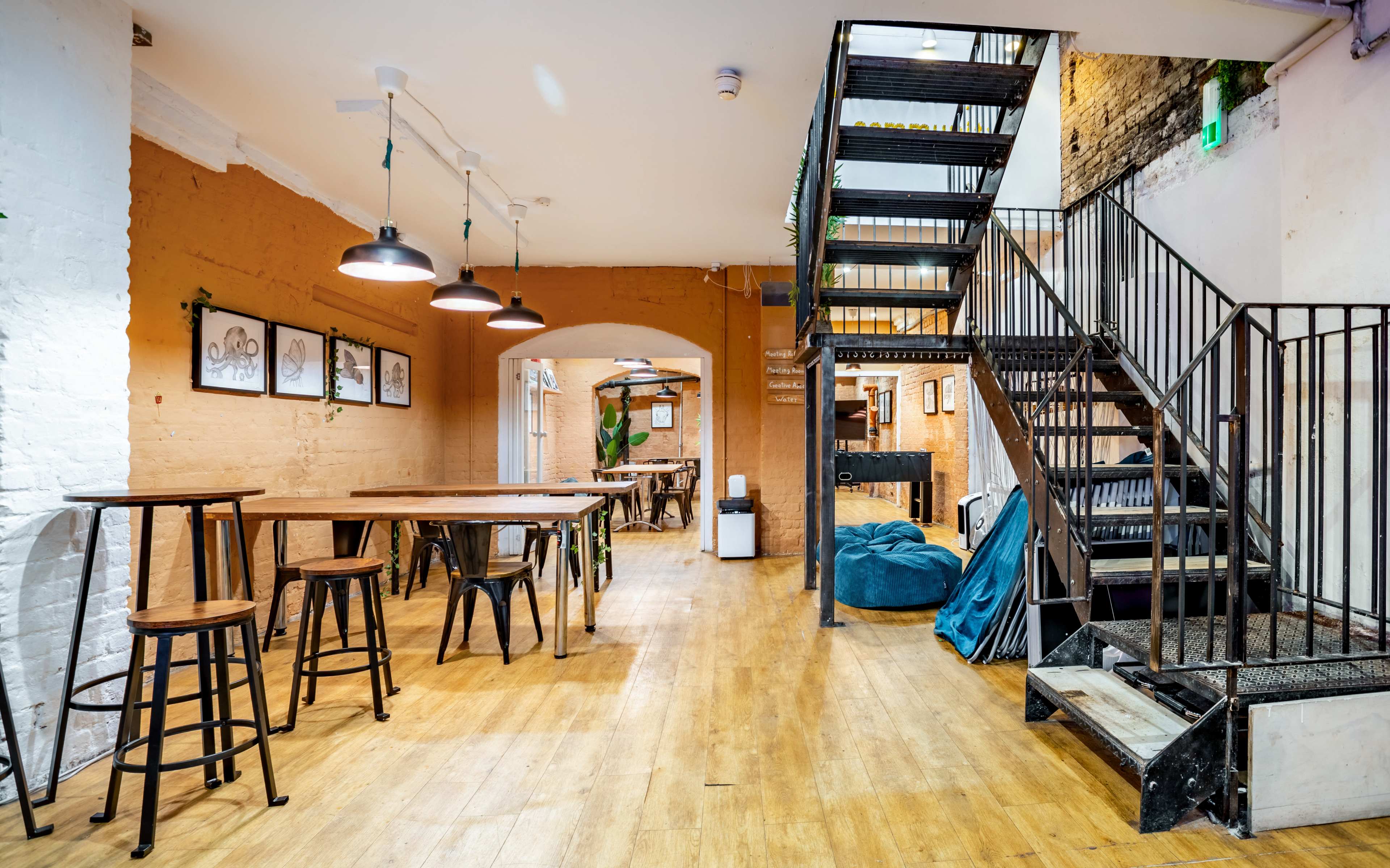 An indoor space with wooden flooring, featuring a staircase, several tables, and a mix of seating options along the walls.