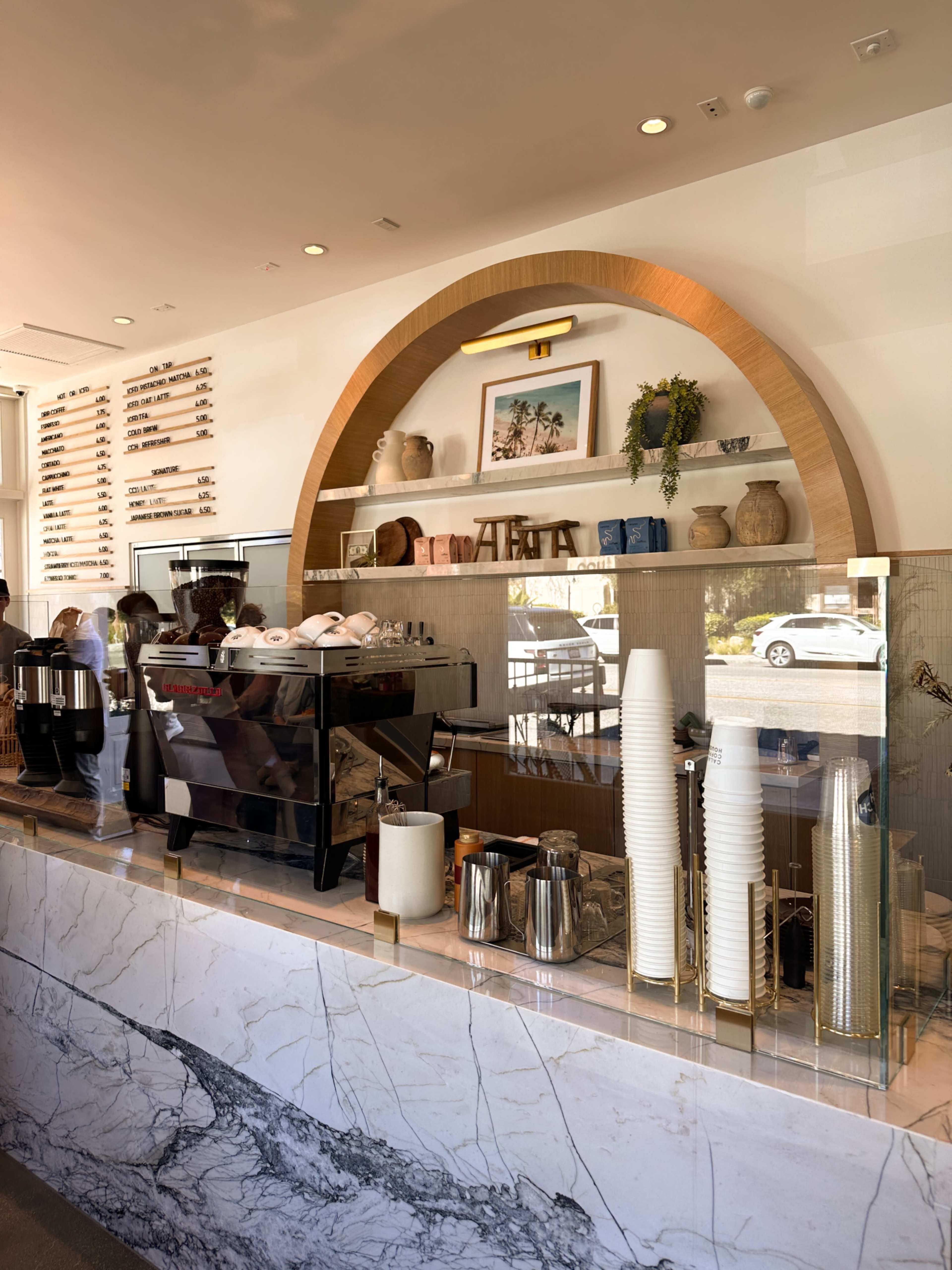 The image shows the interior of a modern coffee shop with a marble counter, a coffee machine, and a display shelf featuring various decorative items and mugs.