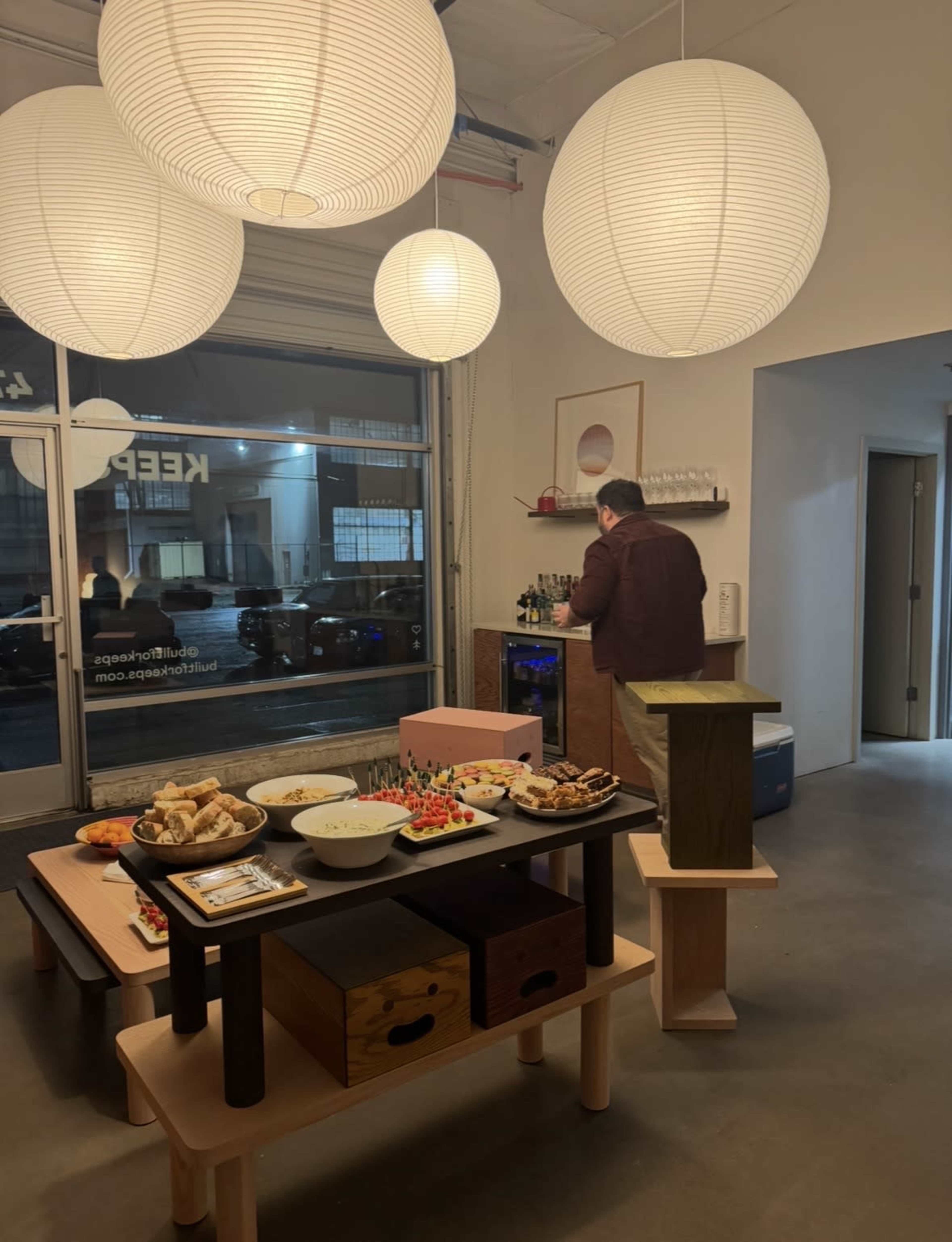 A person stands near a table filled with food, while large paper lanterns hang above in a sparsely decorated room with a glass door showing a city street.
